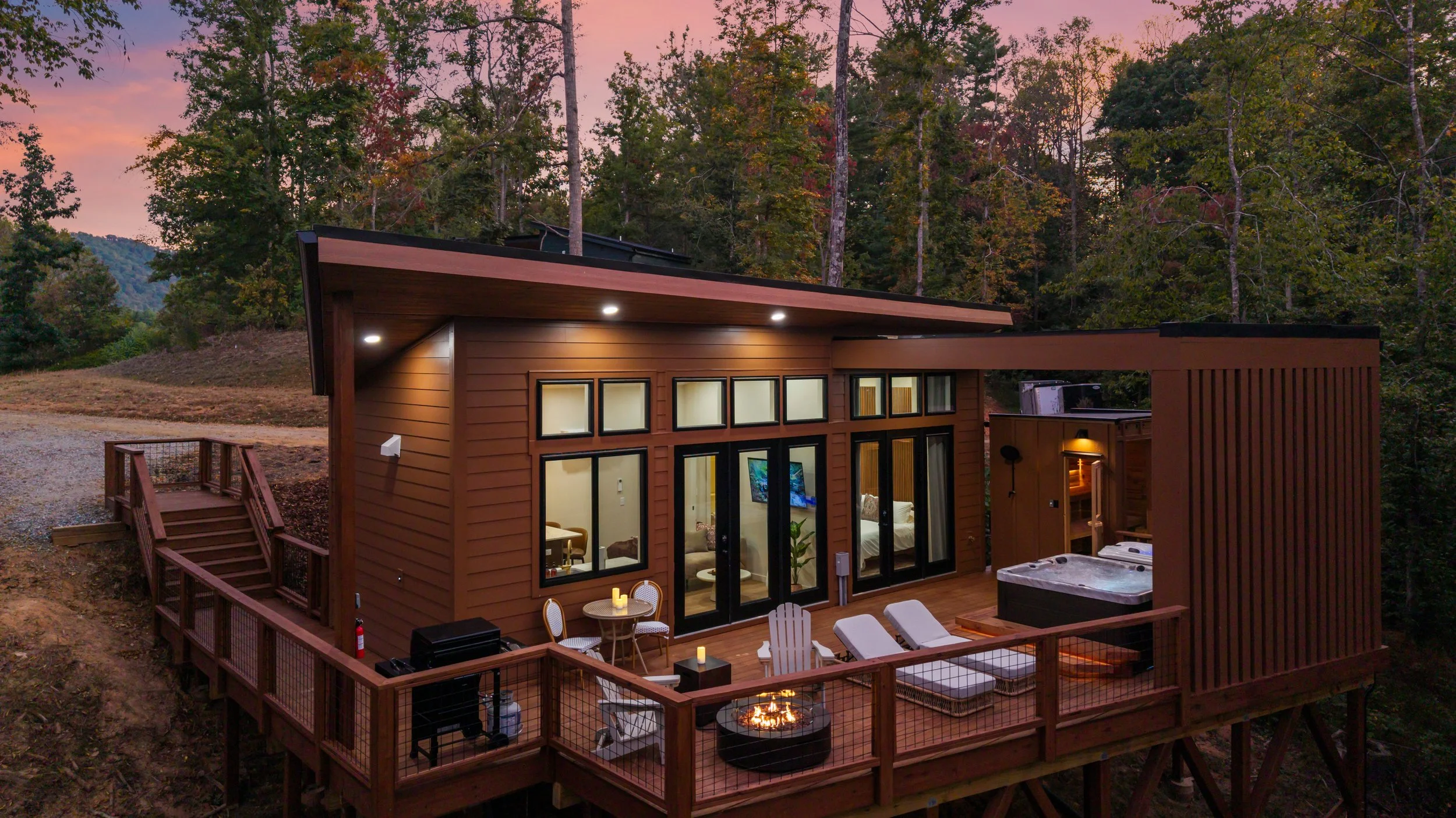 A modern two-story house with large windows and a deck, surrounded by a wooded area, during sunset.