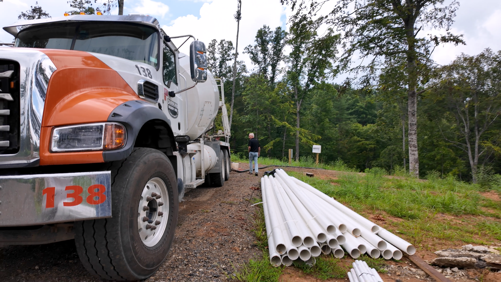 A construction site with a white and orange cement truck labeled '138', parked on a dirt road. Several white pipes are laid out on the ground, and a man in a black shirt and jeans stands nearby. The background features trees and a partly cloudy sky.