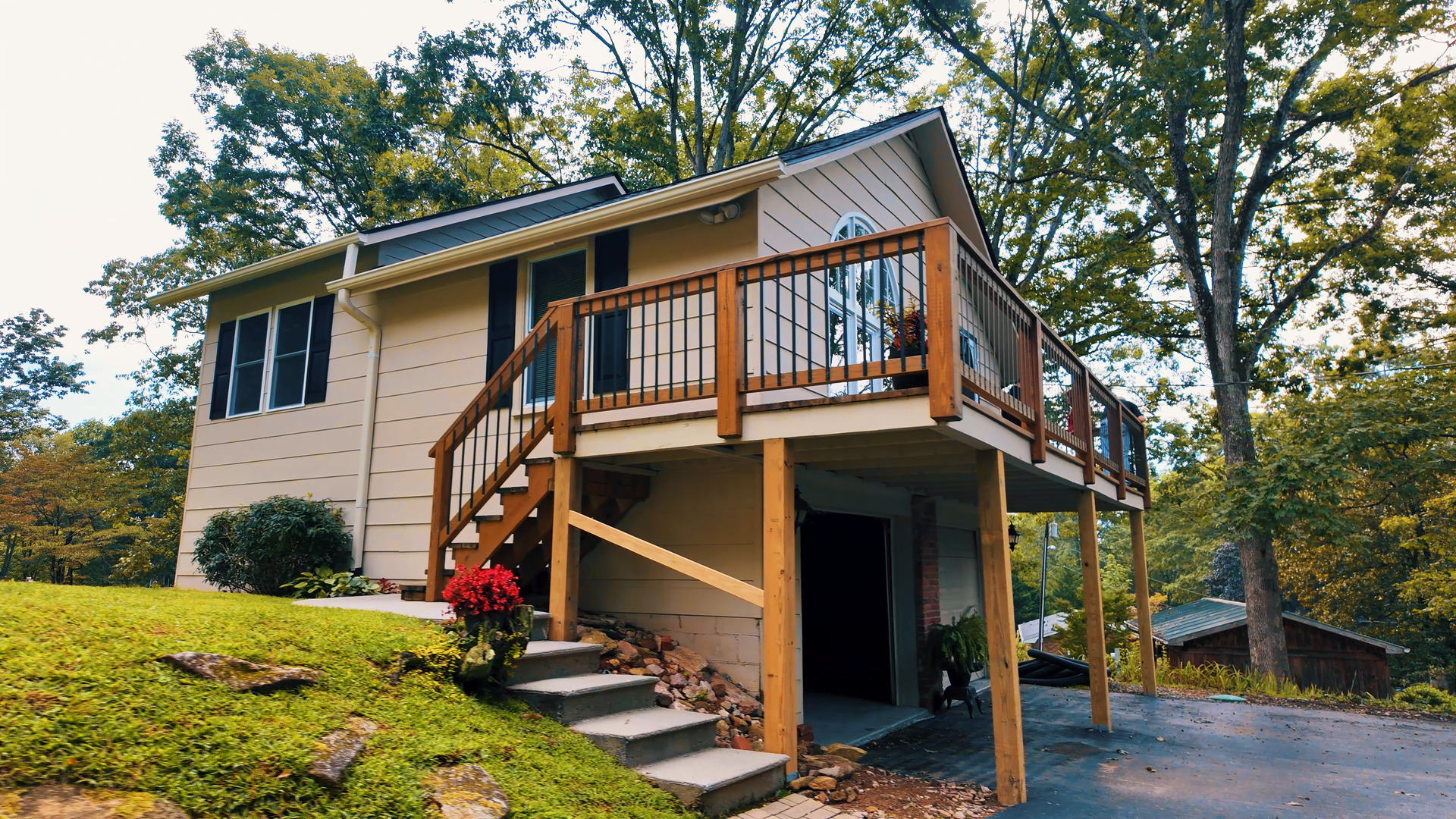 A two-story house with an unfinished elevated deck on the back,visible as a wooden structure with railings and stairs, surrounded by trees and a lawn, with a garage underneath and a paved driveway leading up to it.