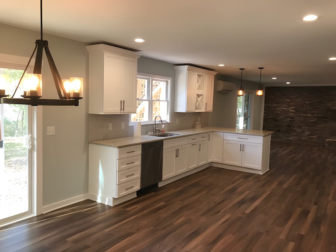 Empty kitchen with white cabinets, beige countertops, and wood flooring, with a stone accent wall in the background and multiple ceiling lights.