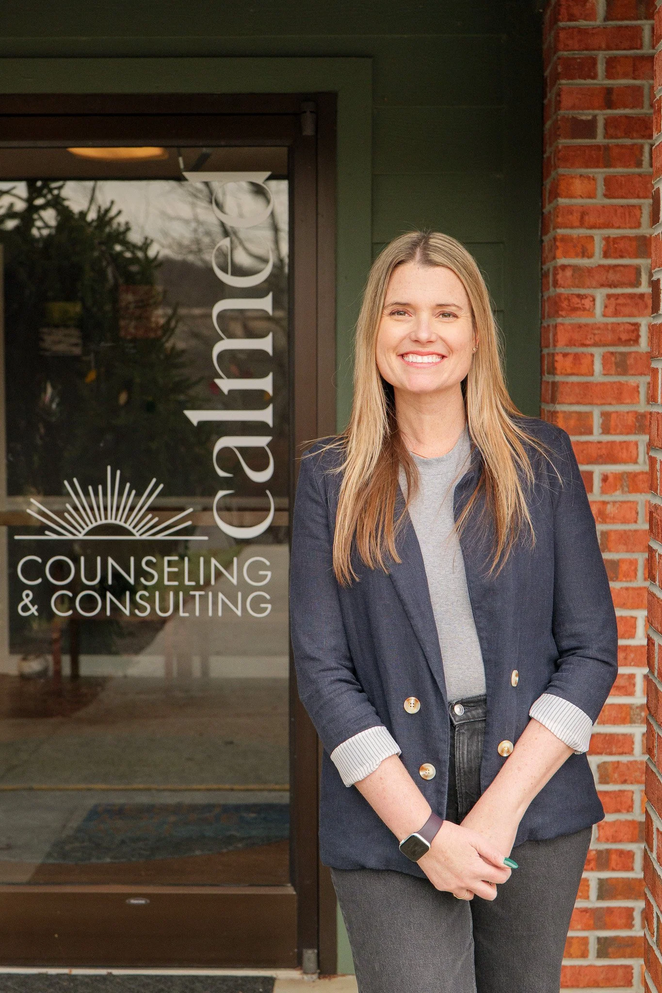 A woman standing outside a counseling and consulting office, smiling, with long blonde hair, wearing a gray top and a navy blazer, next to a brick wall.