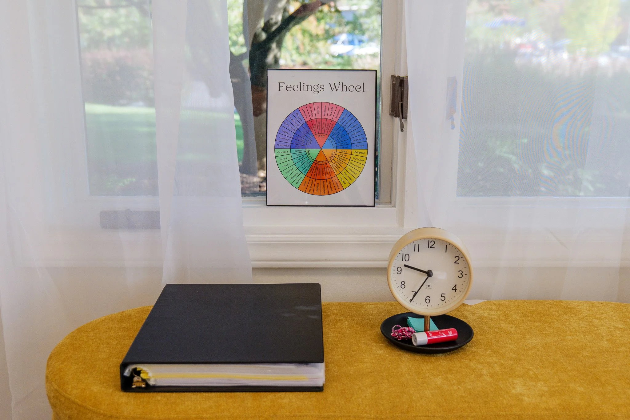 A window with sheer white curtains, a Feelings Wheel chart on the windowsill, a black binder, and a beige clock on a yellow table. The clock shows the time as 9:37.