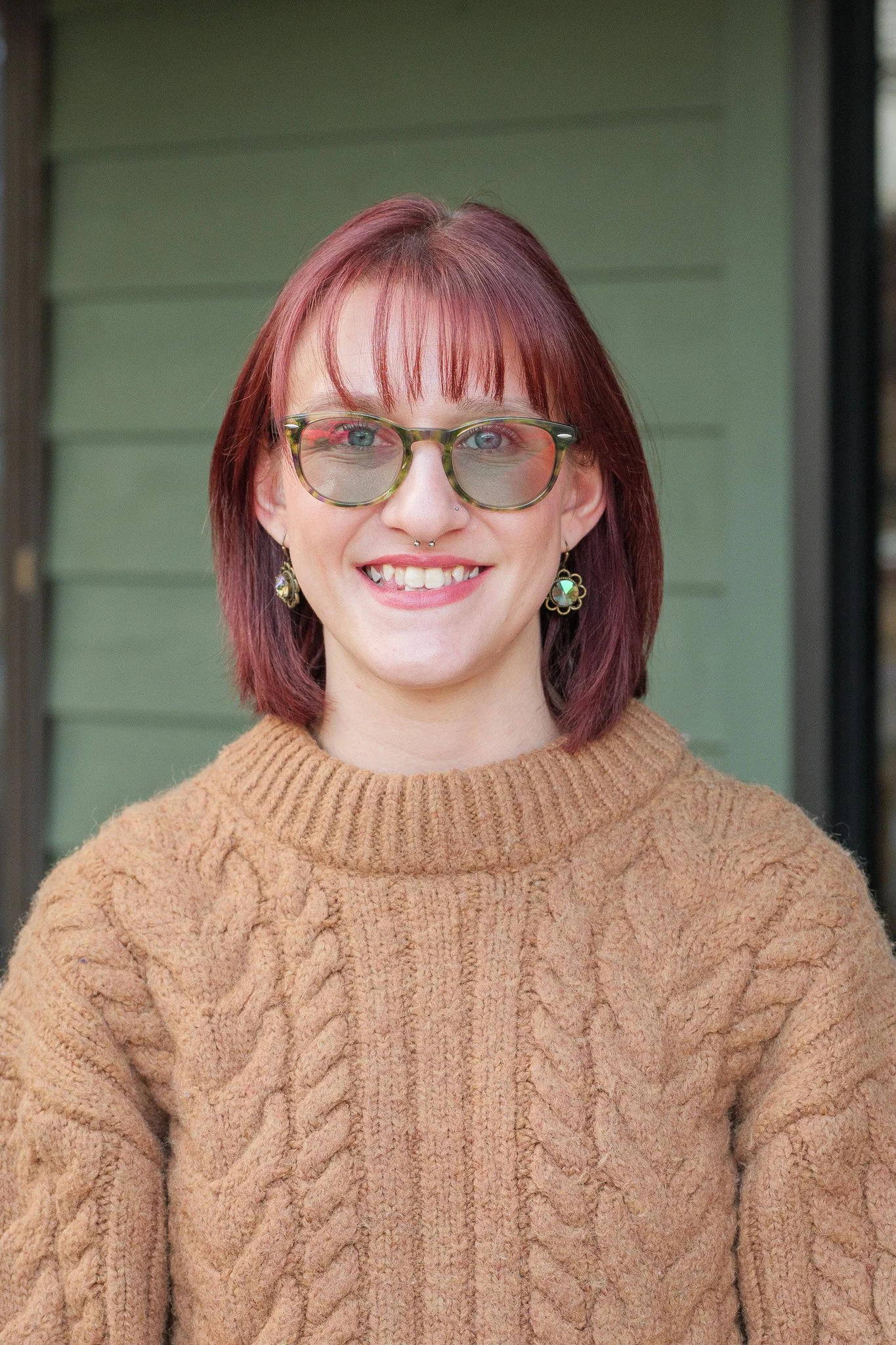A woman with shoulder-length red hair, glasses, and earrings smiling outdoors, wearing a tan knitted sweater.