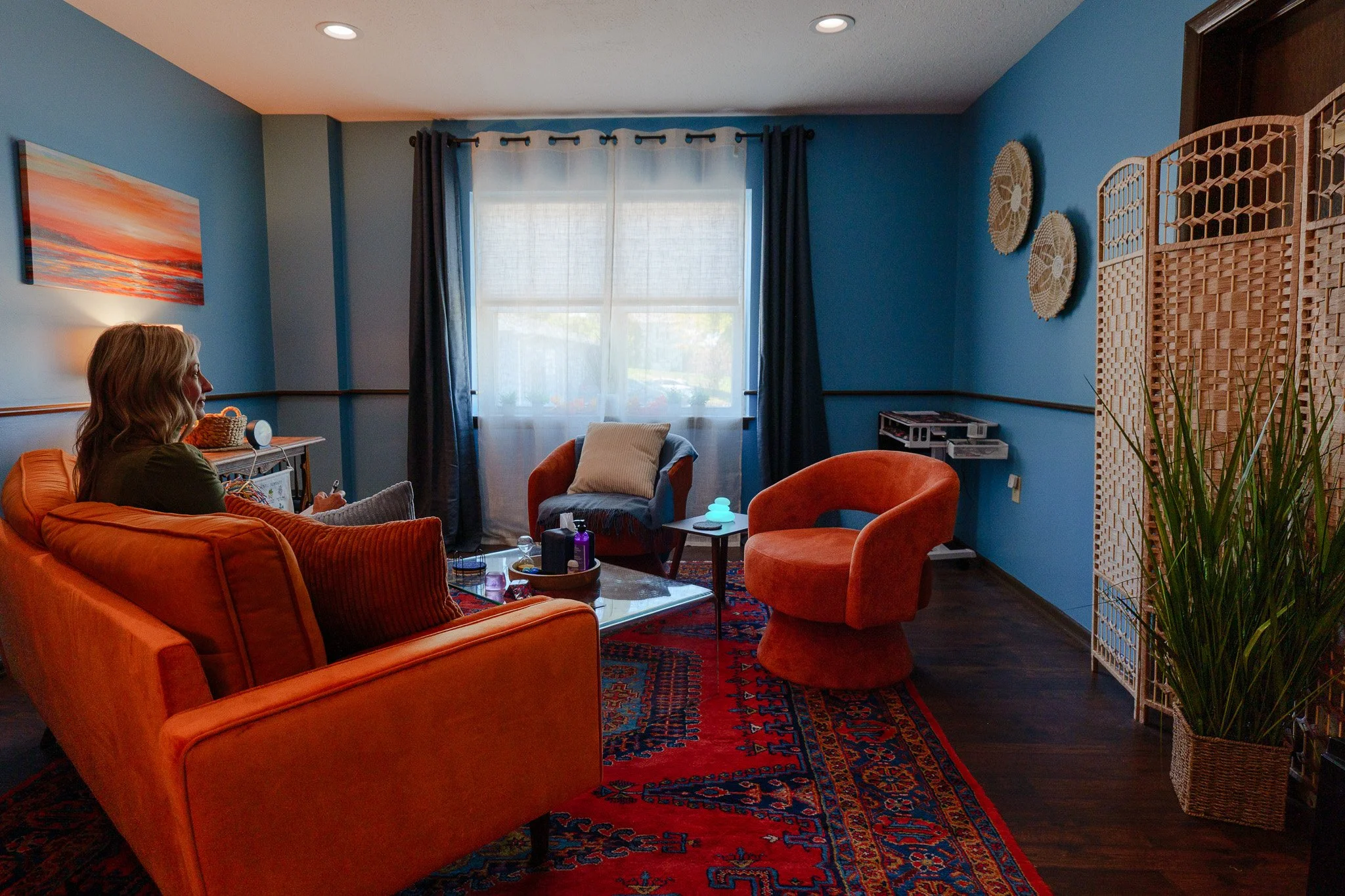 A woman with blonde hair is sitting on an orange sofa in a decorated living room, facing a window with blue curtains. The room has a blue wall, a colorful painting, and a patterned rug, with a table, chairs, a potted plant, and decorative wall art.