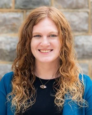 Portrait of a woman with long wavy red hair, smiling, standing outdoors with a stone wall background.