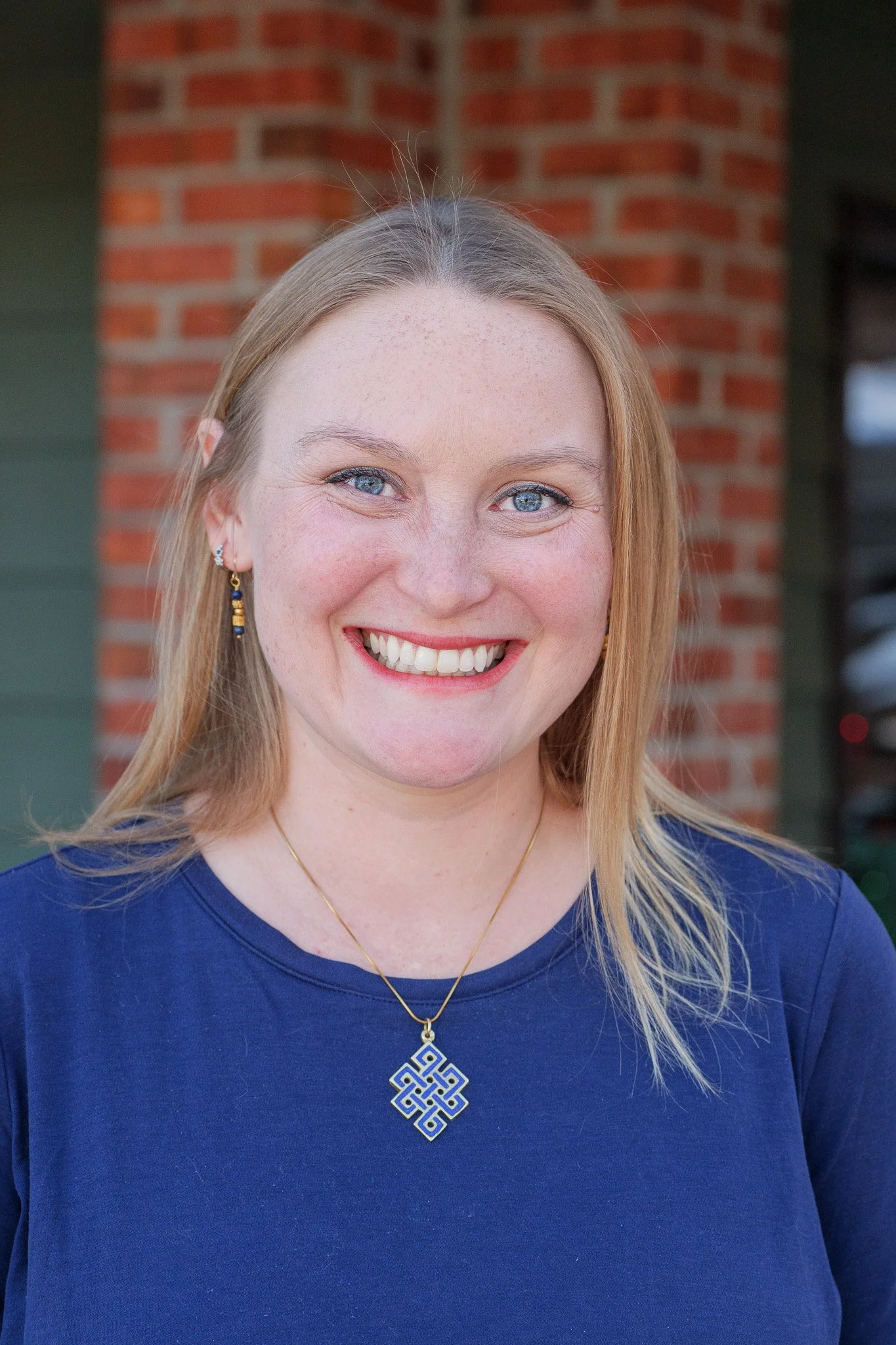 Close-up of smiling woman with blue eyes and blonde hair, wearing a blue top, earrings, and a necklace with a geometric pendant, standing outdoors in front of a brick wall.