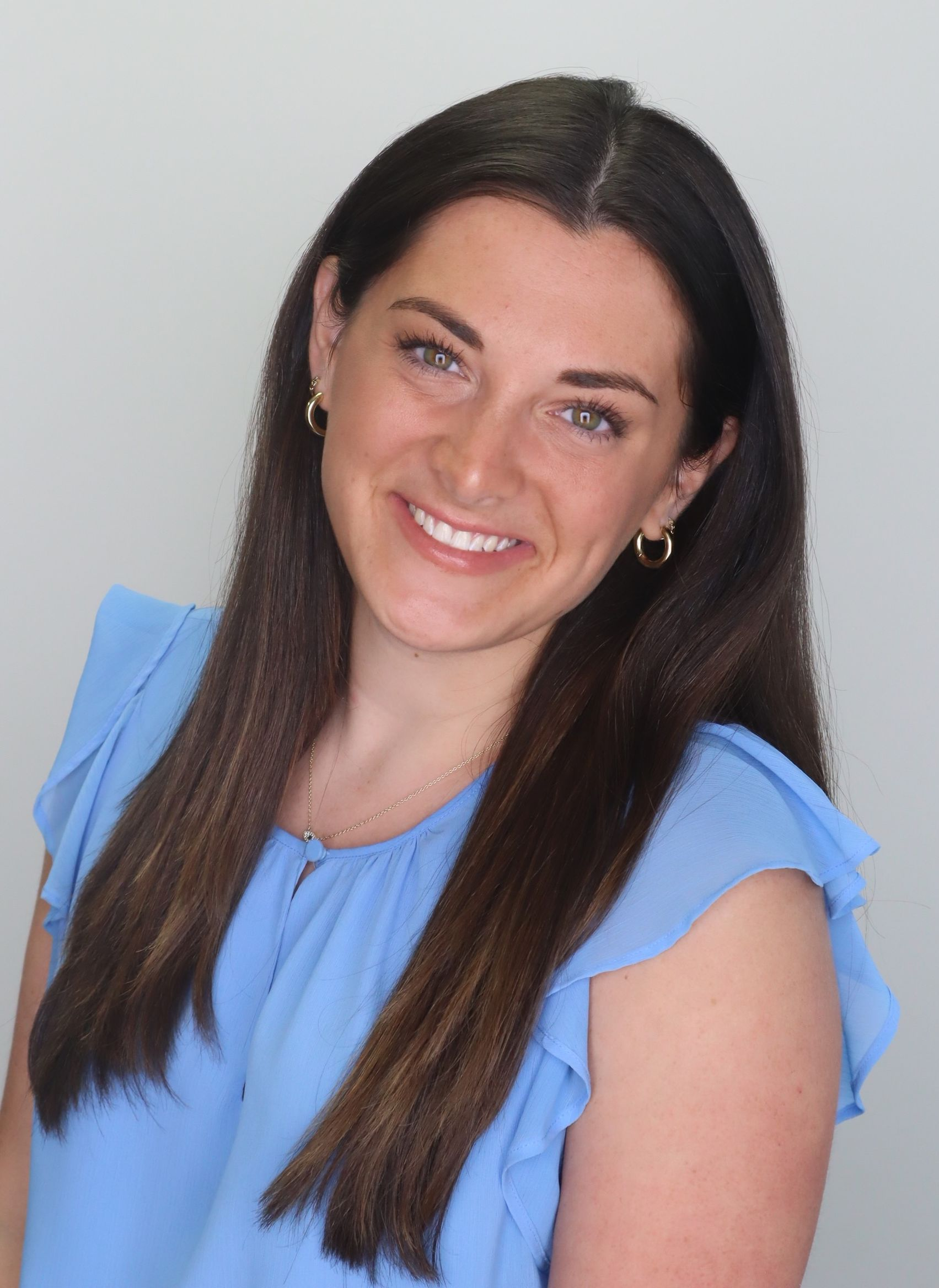 A woman with long brown hair wearing a light blue sleeveless top, gold hoop earrings, and a necklace, smiling at the camera against a plain background.