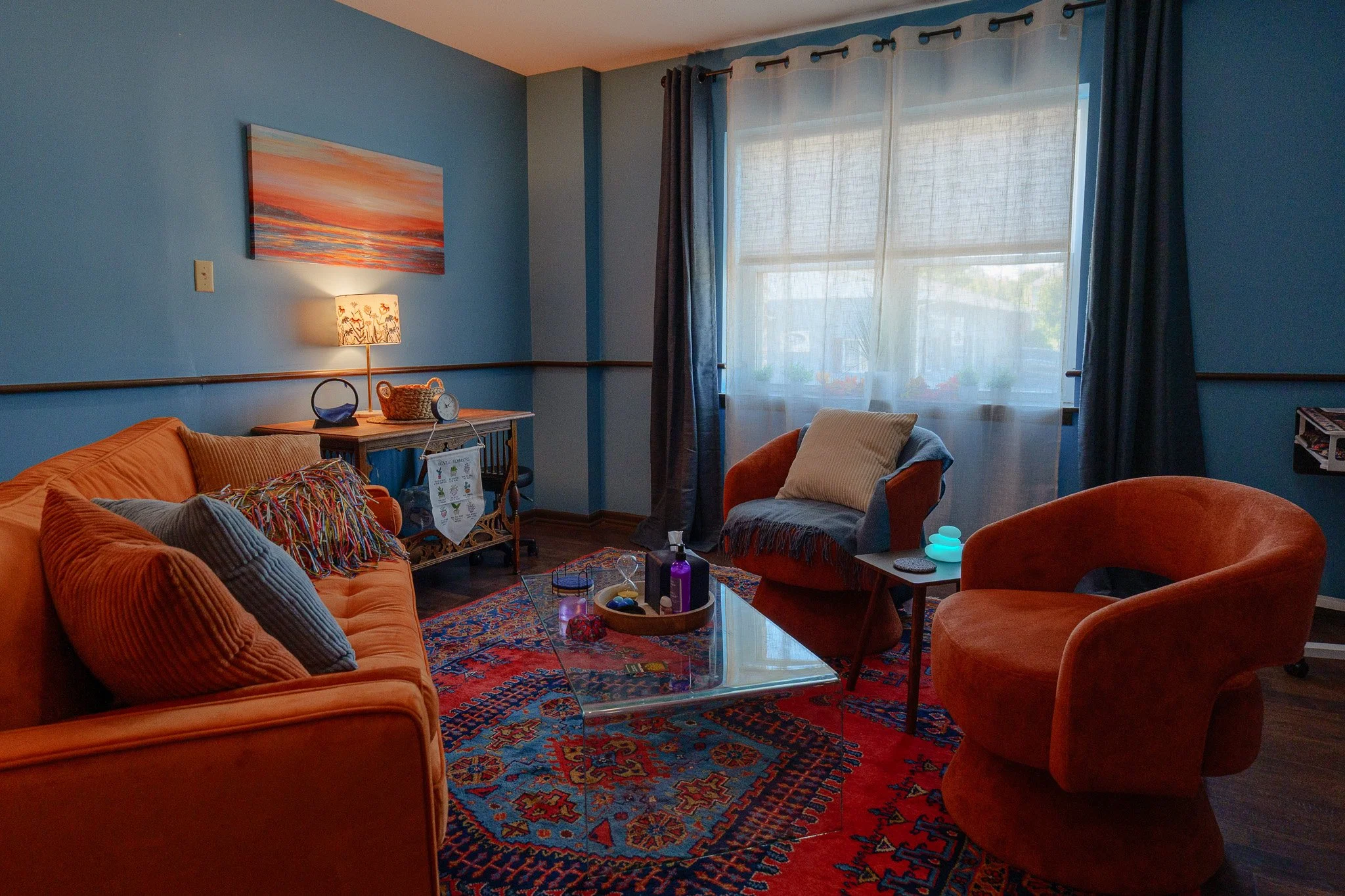 Living room with blue walls, orange sofa with pillows, a colorful rug, a glass coffee table, a patterned armchair with a pillow, large window with curtains, a side table, and decorative lamps.