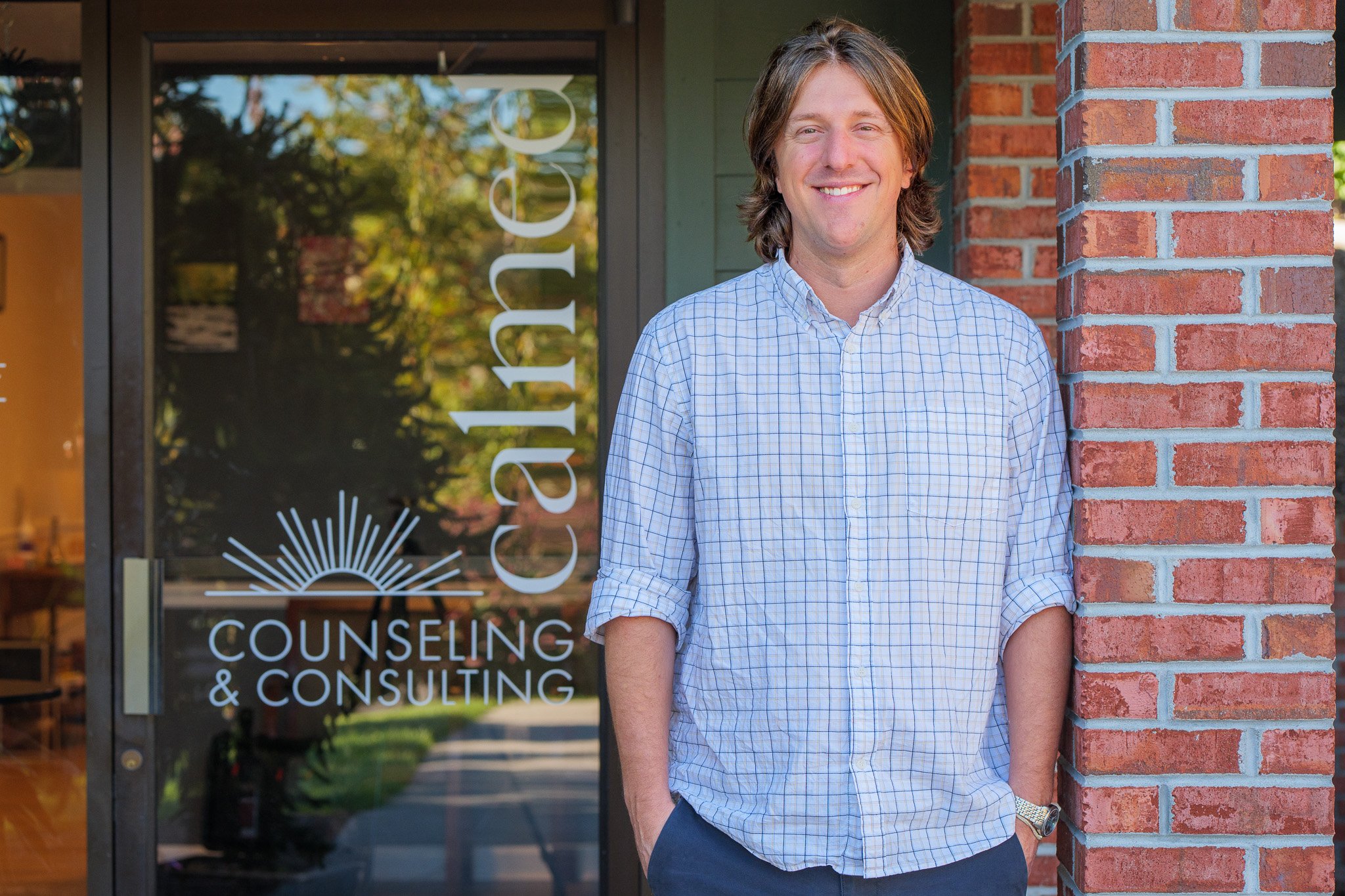 A man smiling and standing outside counseling and consulting office, leaning against a brick wall next to the glass door.