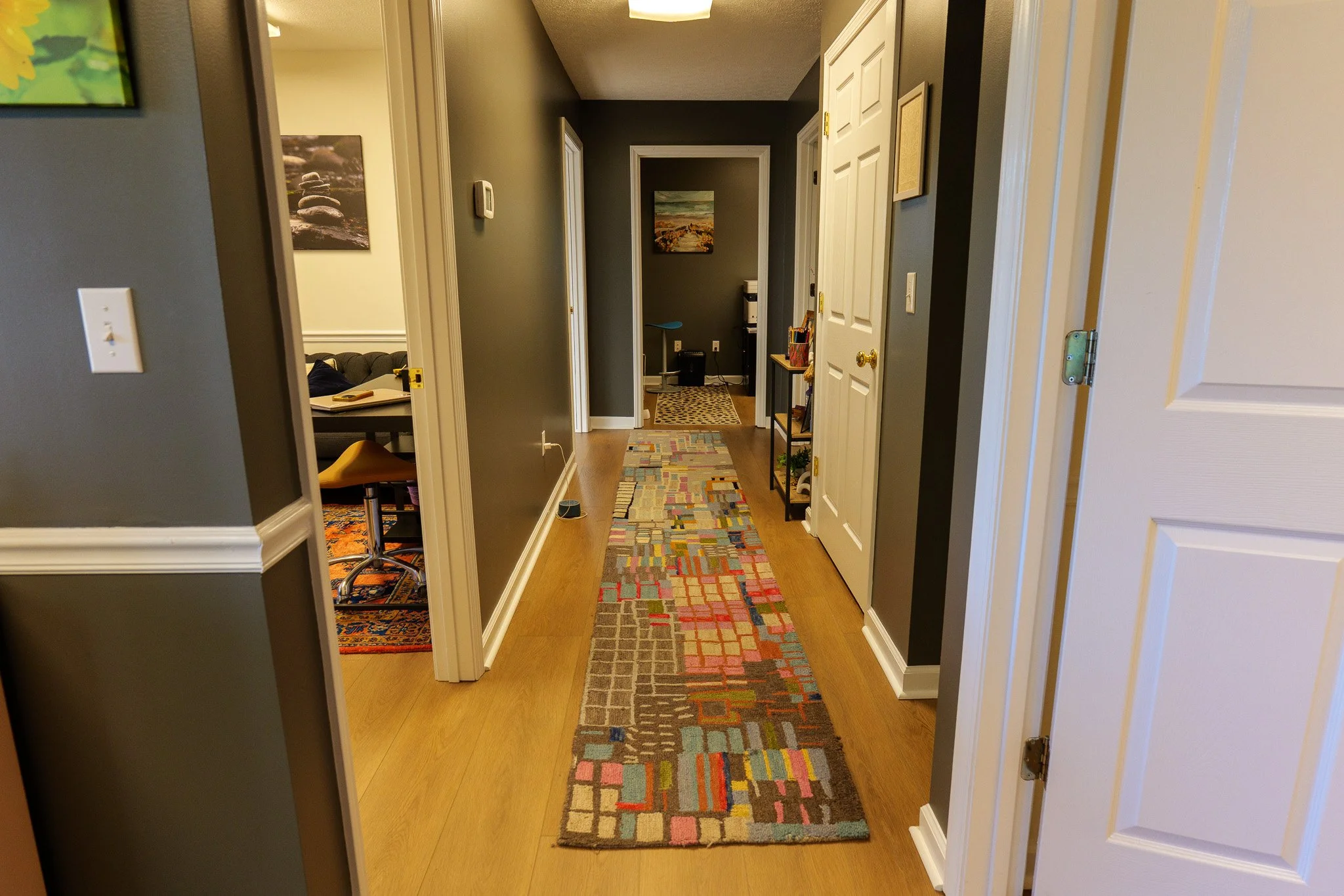 A hallway with colorful geometric patterned runner rug on wooden floor, gray walls, white trim, and multiple doorways leading to rooms with artwork on the walls.