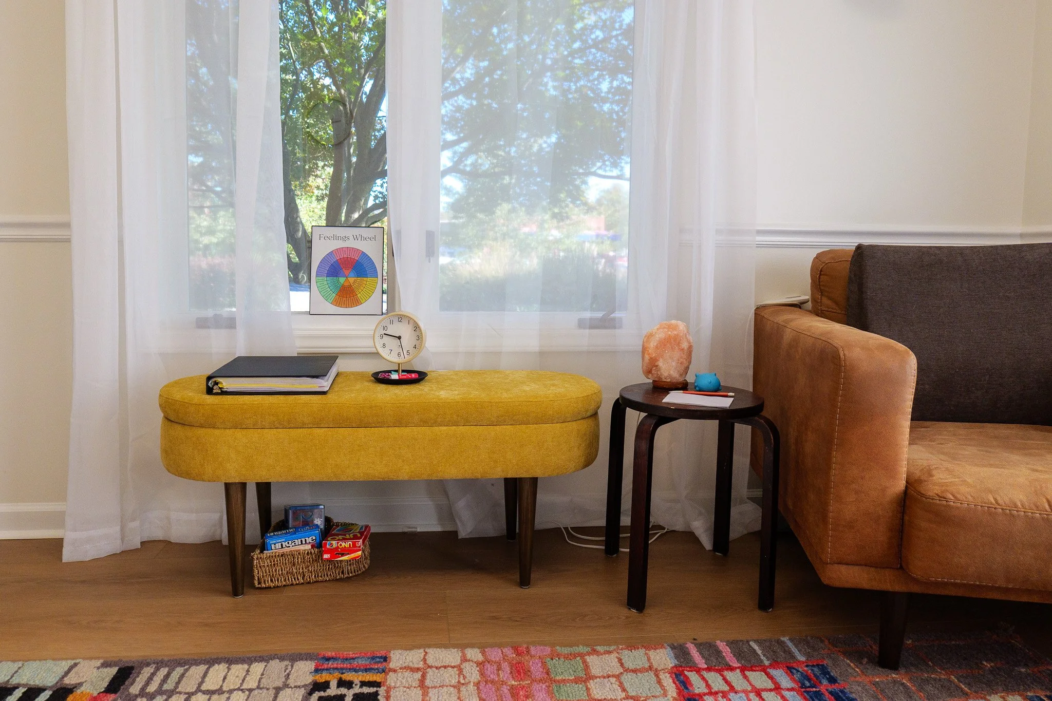 A living room corner with a yellow cushioned bench, a small dark wooden side table holding salt lamp, salt rock, a blue figurine, and a notebook. Behind the bench is a window with white sheer curtains showing green trees outside. A framed picture titled 'Feelings Wheel' and a clock are on the bench. Below the bench is a wicker basket with board games. Part of a tan and dark gray couch is on the right, and a colorful patterned rug is on the wooden floor.