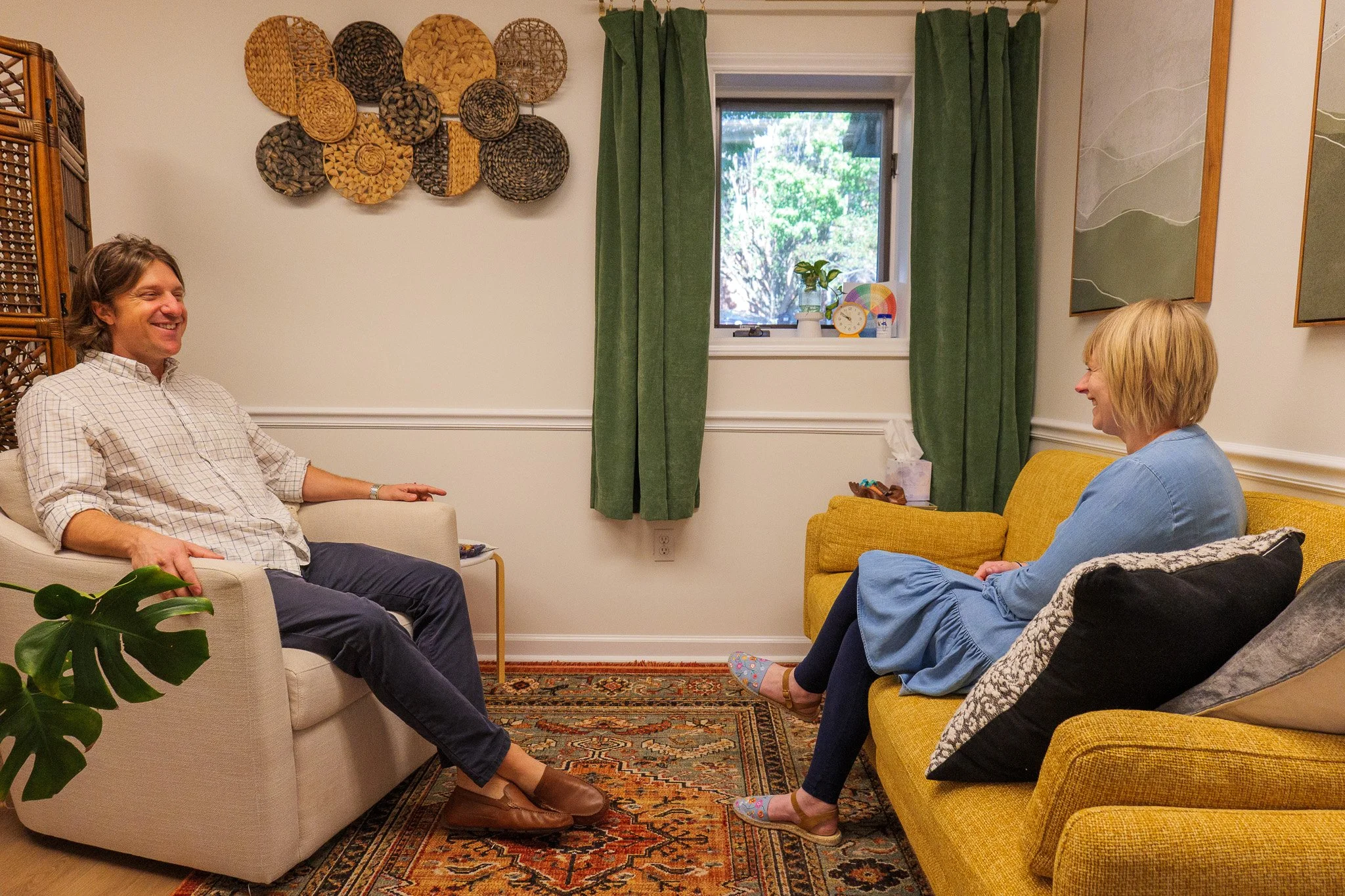 A man and woman sitting on separate couches in a living room, engaged in conversation and smiling. The man is on a beige couch, wearing a checkered shirt and dark pants. The woman is on a yellow couch, wearing a blue dress and slippers. There are green curtains, wall art, and decorative woven baskets on the wall.