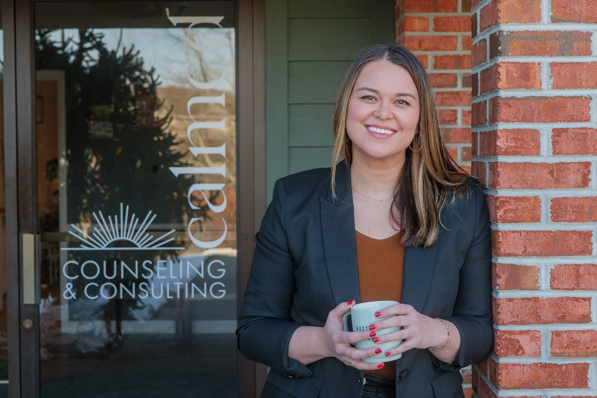 A woman smiling and holding a coffee mug outside a counseling and consulting office, with a glass door displaying the business name.