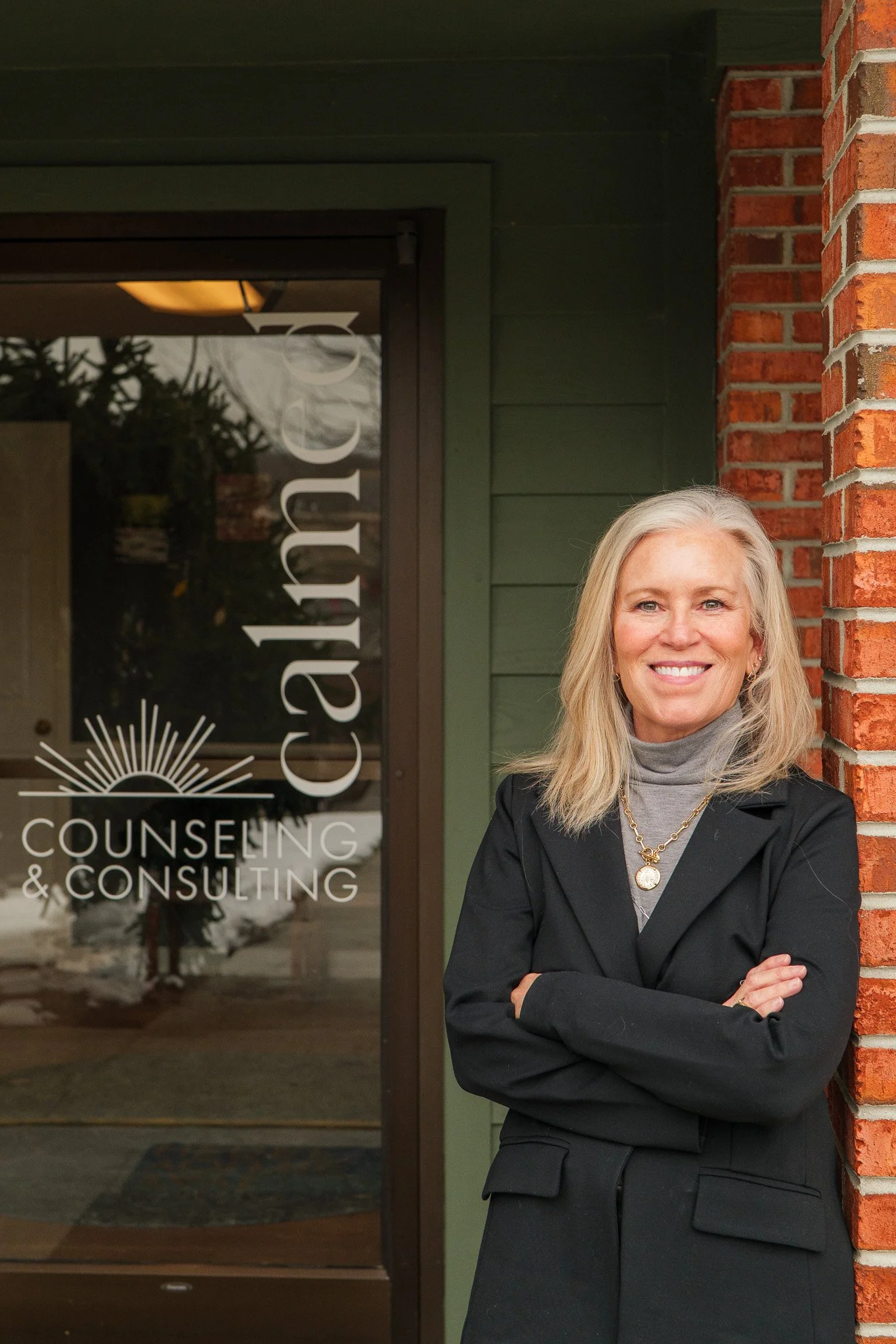A woman with blonde hair wearing a black blazer and gray turtleneck standing outside a counseling and consulting office, smiling with arms crossed. The office window behind her displays the business name.