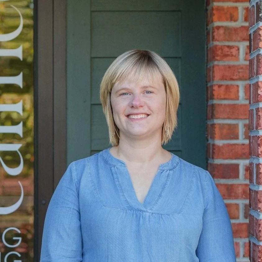 Portrait of a young woman with blonde hair, smiling, standing in front of a green building and a brick wall.