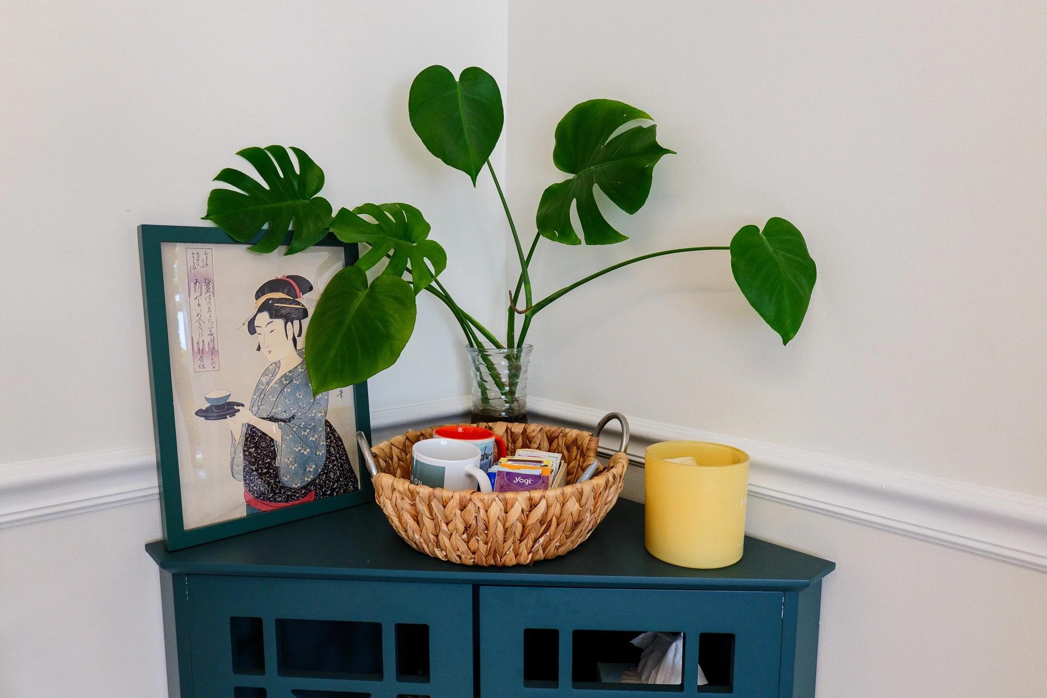 A teal console table with a potted Monstera plant, a framed Japanese-style artwork, a woven basket holding mugs and tea bags, and a yellow candle.