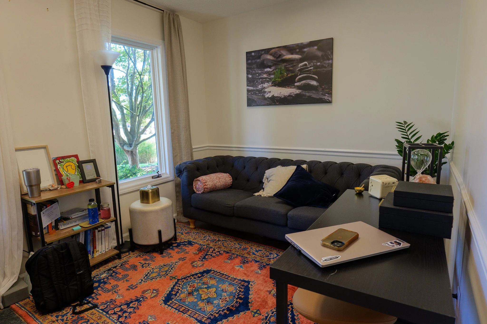 A cozy living room with a dark gray tufted sofa, a colorful orange and blue patterned rug, a tall window with white curtains, and a small bookshelf with books and decorative items. There is a floor lamp next to the window, a small off-white ottoman with a gold decorative piece on it, and a black desk with a laptop, a smartphone, and a stack of black boxes. A sofa pillow and a bolster pillow are on the sofa, and a framed artwork of stacked stones is on the cream-colored wall. The room also has a green plant with a glass terrarium and an hourglass on a side table.