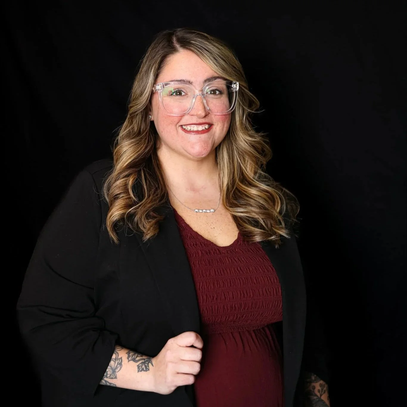 A woman with wavy, shoulder-length hair, wearing glasses, a black blazer, and a dark red top, poses with a smile against a black background.