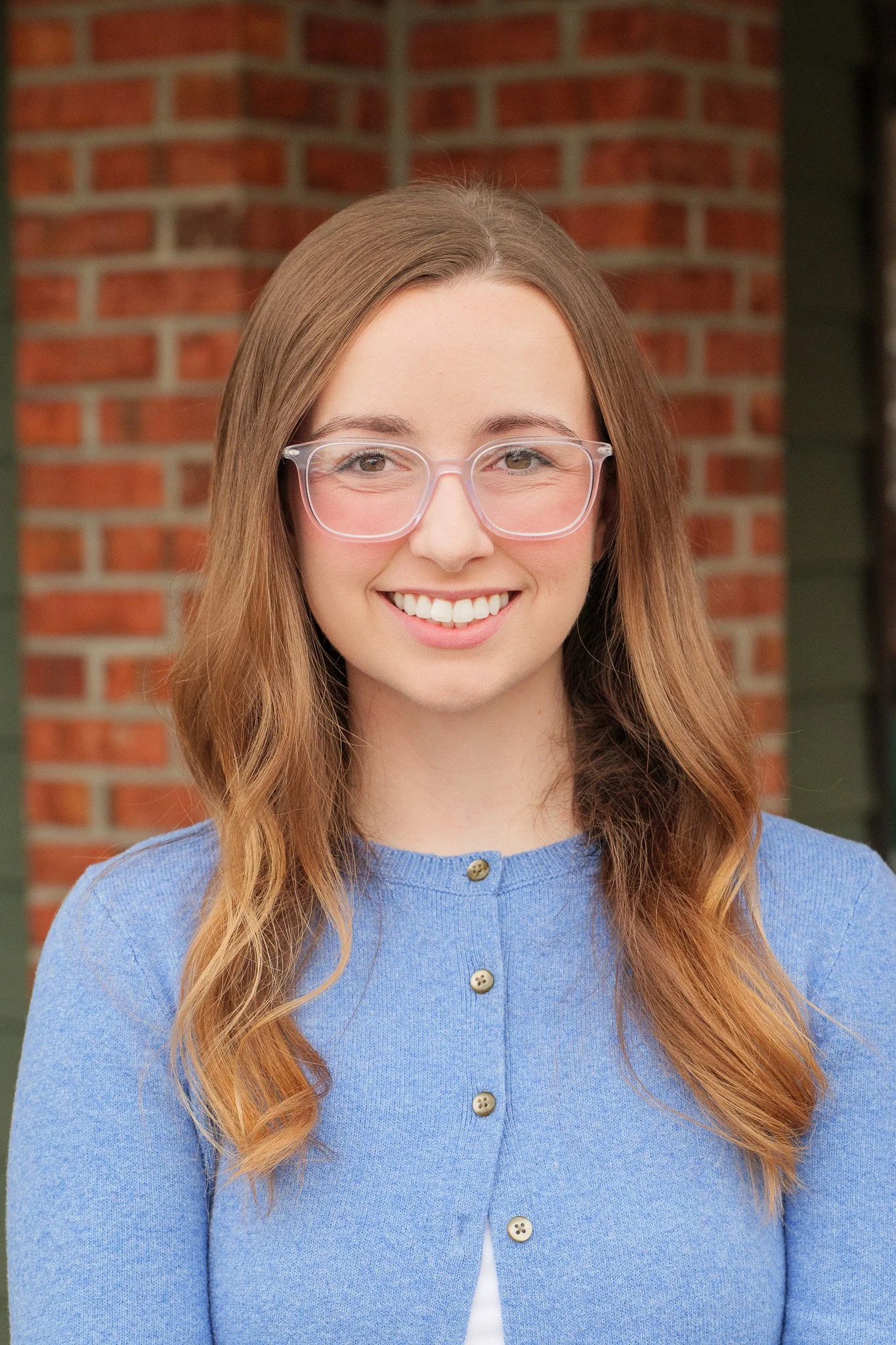 A young woman with long, wavy auburn hair, wearing clear glasses and a light blue cardigan, smiling outdoors with a brick wall in the background.