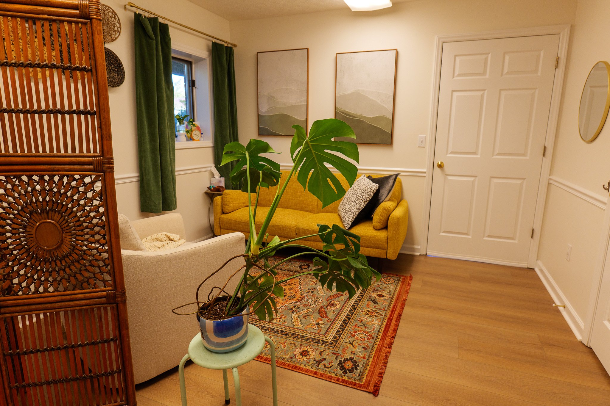 A cozy living room with a yellow sofa, cream armchair, potted monstera plant, green curtains, wall art, and a small table, on a decorative rug.