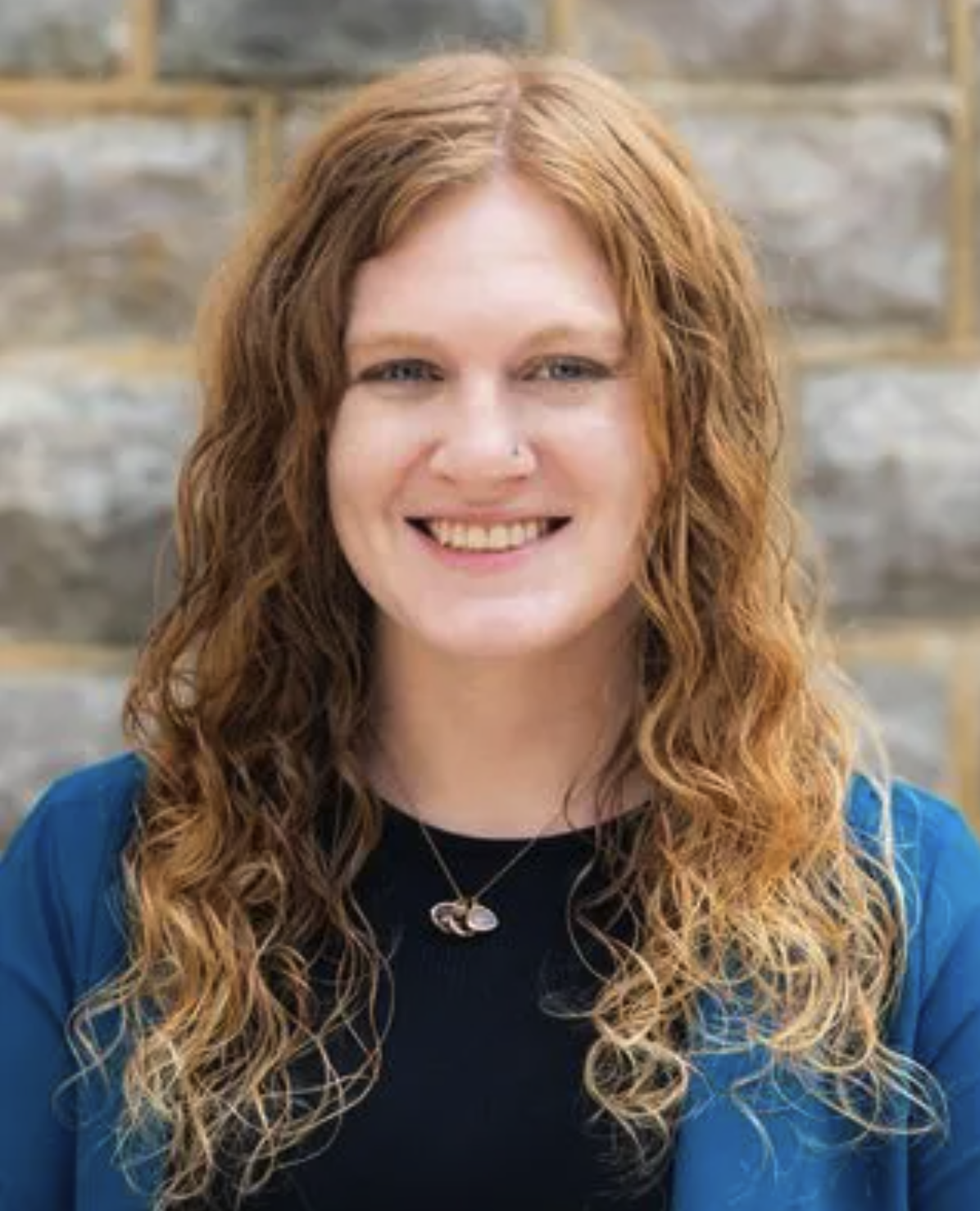 A woman with curly red hair, smiling, wearing a black top and a blue jacket, with a stone wall background.