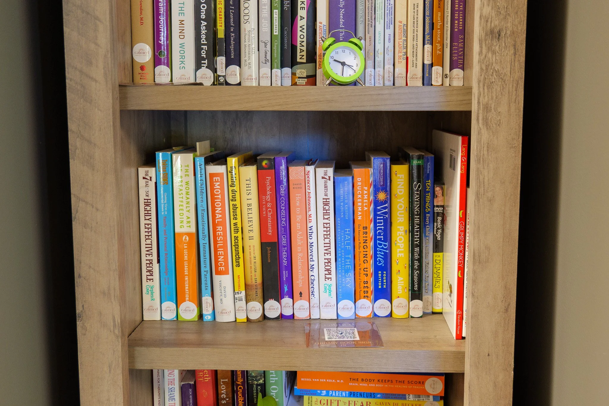 A wooden bookshelf with three shelves filled with various books. The top shelf has a small green alarm clock. The middle shelf contains a variety of colorful books, mostly on psychology, self-help, and health topics. The bottom shelf also has books on health and wellness.