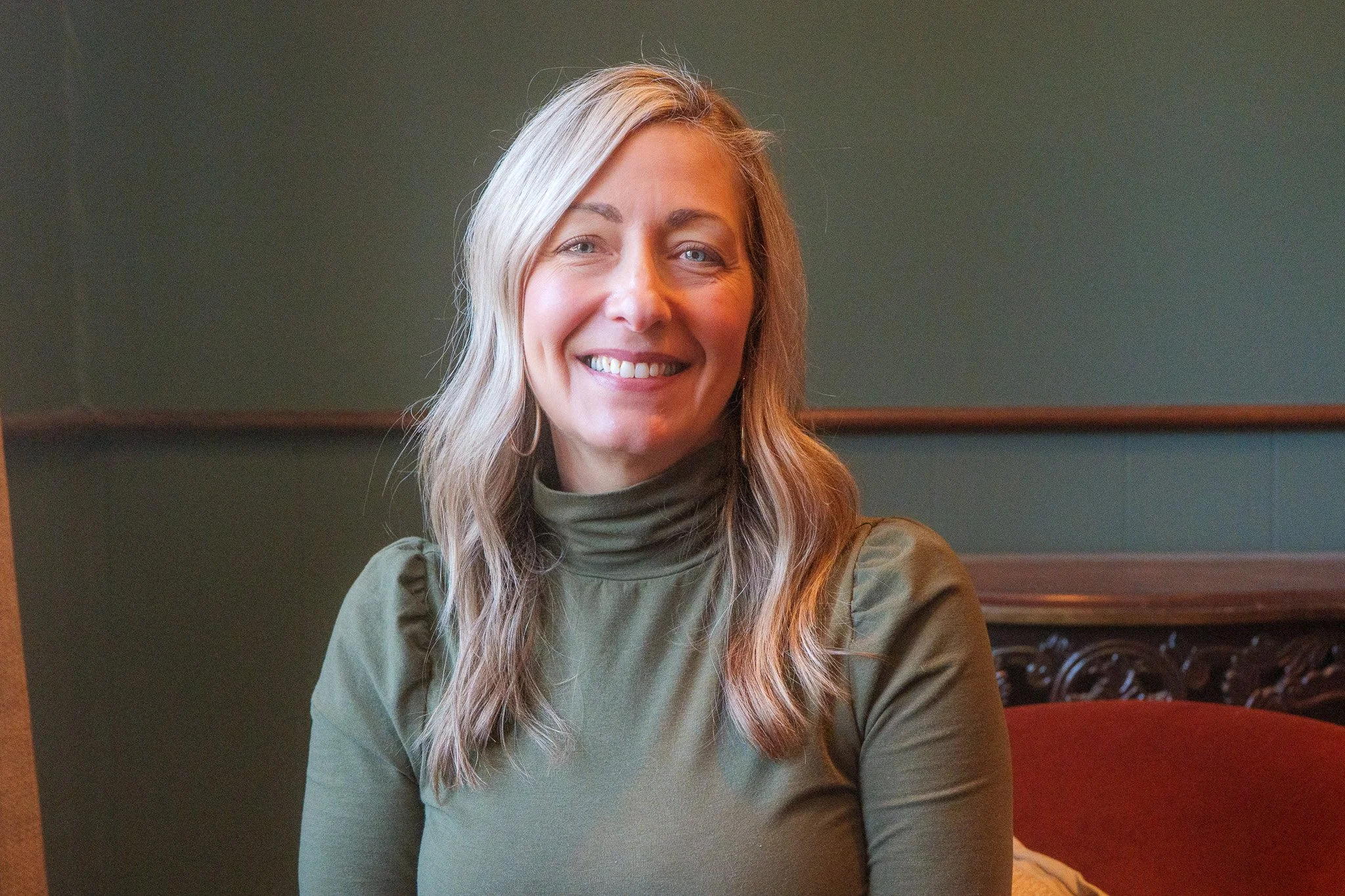 A woman with blonde hair wearing a green turtleneck sweater, smiling and sitting indoors against a green wall.