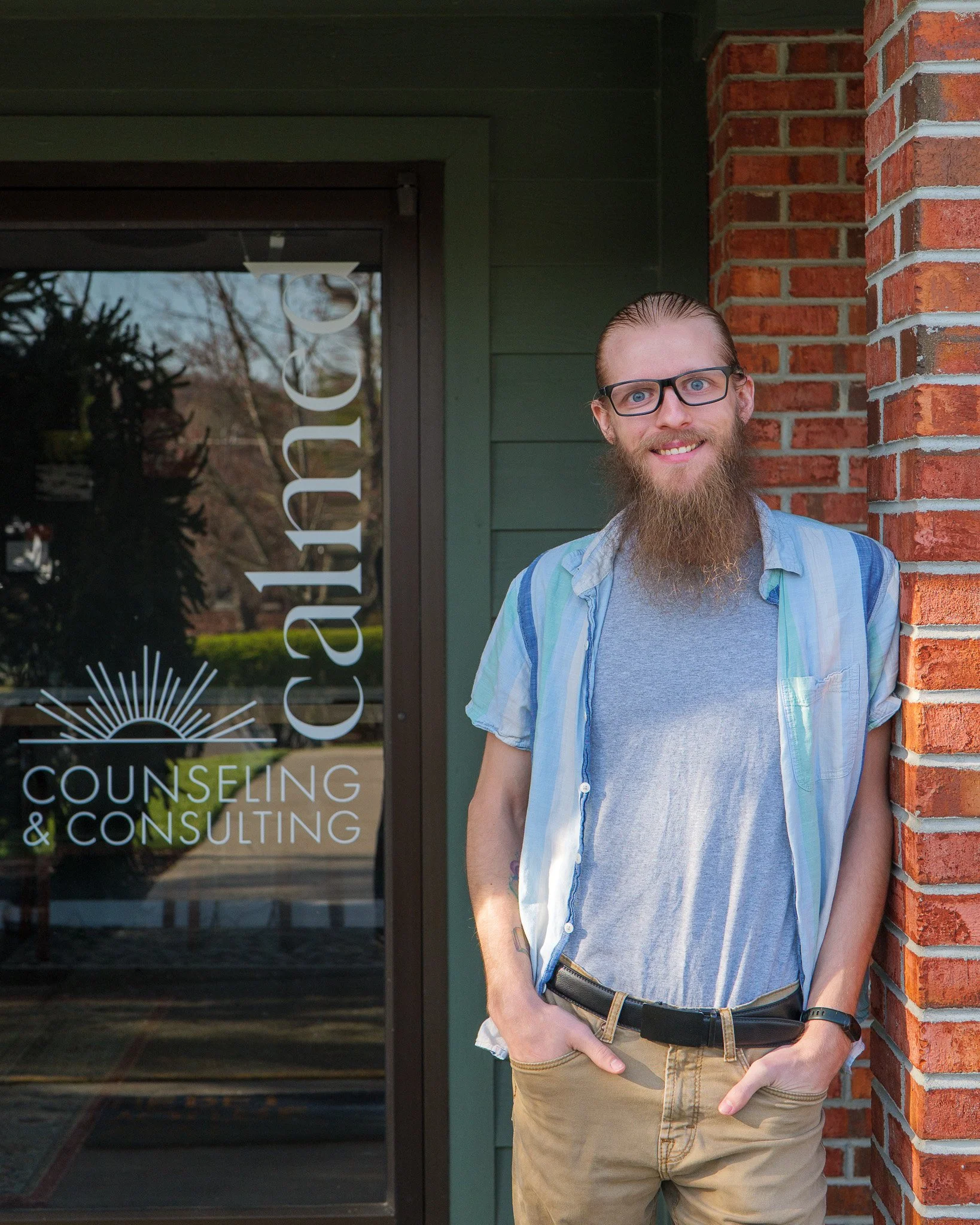 A young man with glasses, a beard, and long hair standing outside Counseling & Consulting office, smiling and leaning against a red brick wall.