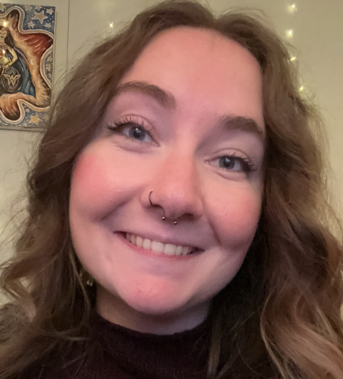Close-up of a smiling woman with wavy light brown hair, light skin, and blue eyes, wearing a septum piercing, sitting indoors with a Wonder Woman poster and yellow lights in the background.