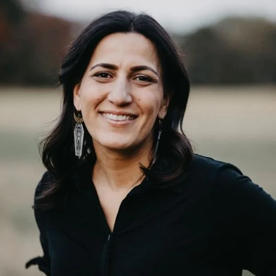 Woman with dark hair smiling outdoors, wearing a black top and earrings.
