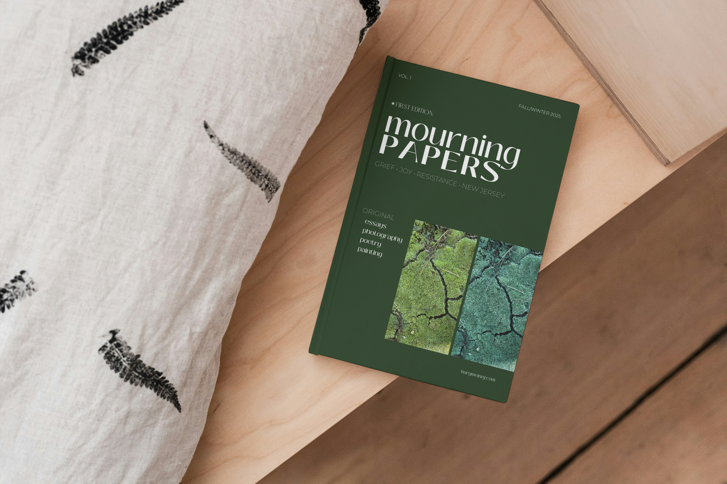 A green magazine titled 'Mourning Papers' resting on a light wooden surface, with a beige fabric with black fern prints partially underneath.
