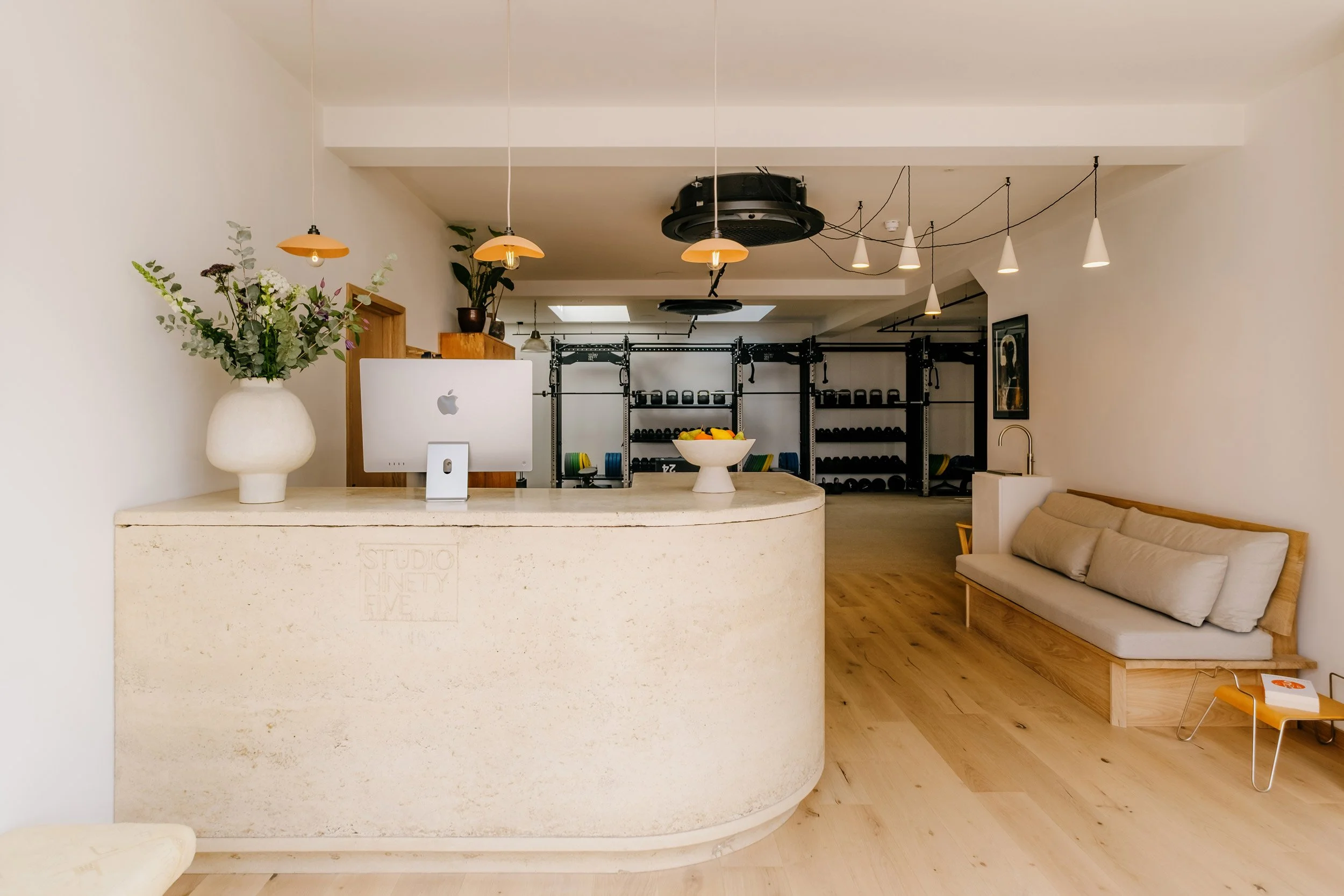 Reception area with a curved beige marble counter, a white iMac computer, a vase with green and purple flowers, and a small framed photo. Behind the counter is a fitness area with weights and exercise equipment. To the right, there is a beige padded
