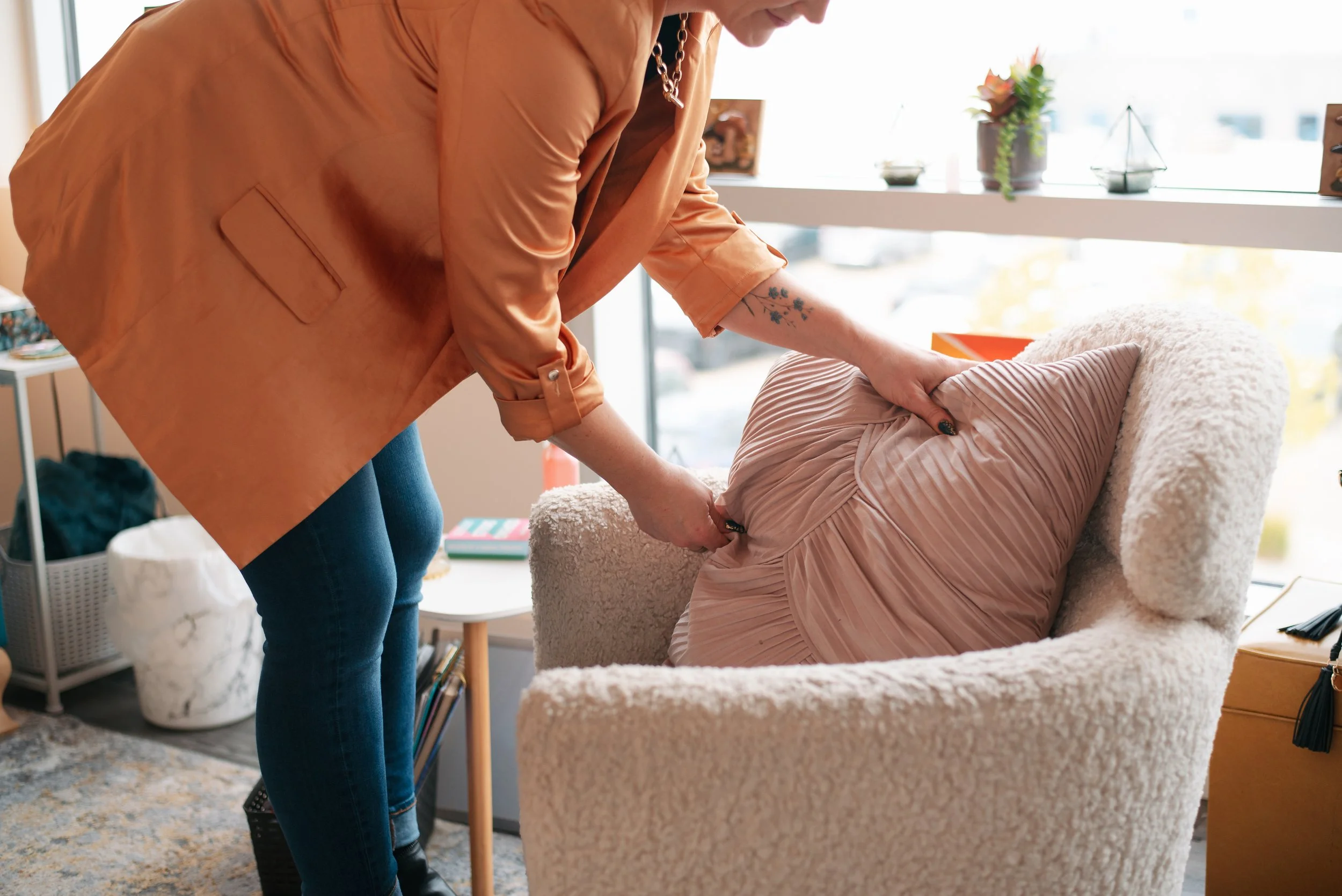 Person in an orange blouse and blue jeans adjusts a pink, pleated, slip dress on a cozy beige armchair in a bright room with a large window and decorated shelf.