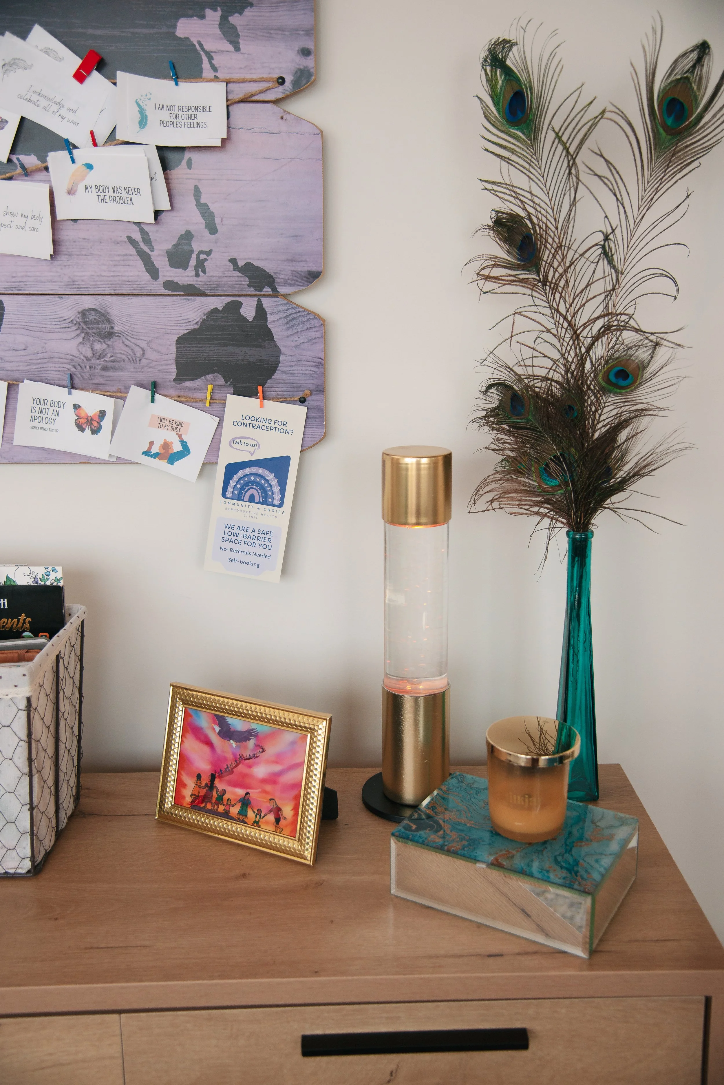 A wooden dresser with a framed photo of people holding hands in front of a sunset sky, a gold candle holder, and a decorative box with a marble pattern. Next to the dresser is a tall blue glass vase with peacock feathers, and a gold and transparent d