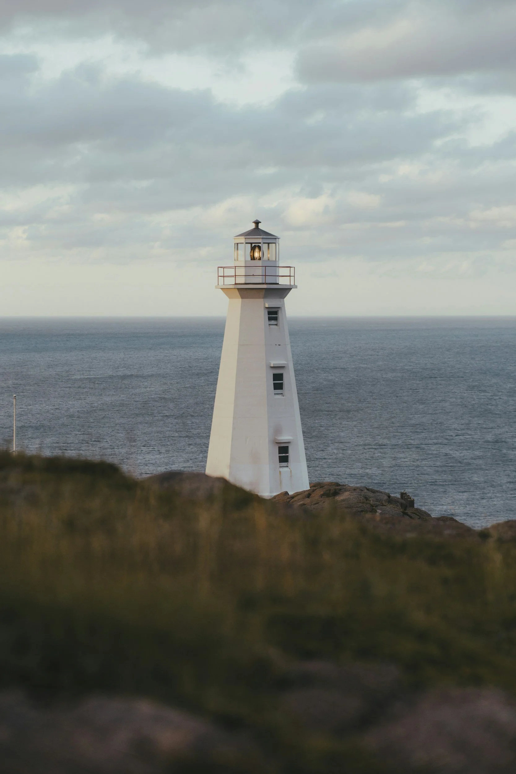 A white lighthouse standing on rocky terrain by the ocean with cloudy sky overhead.