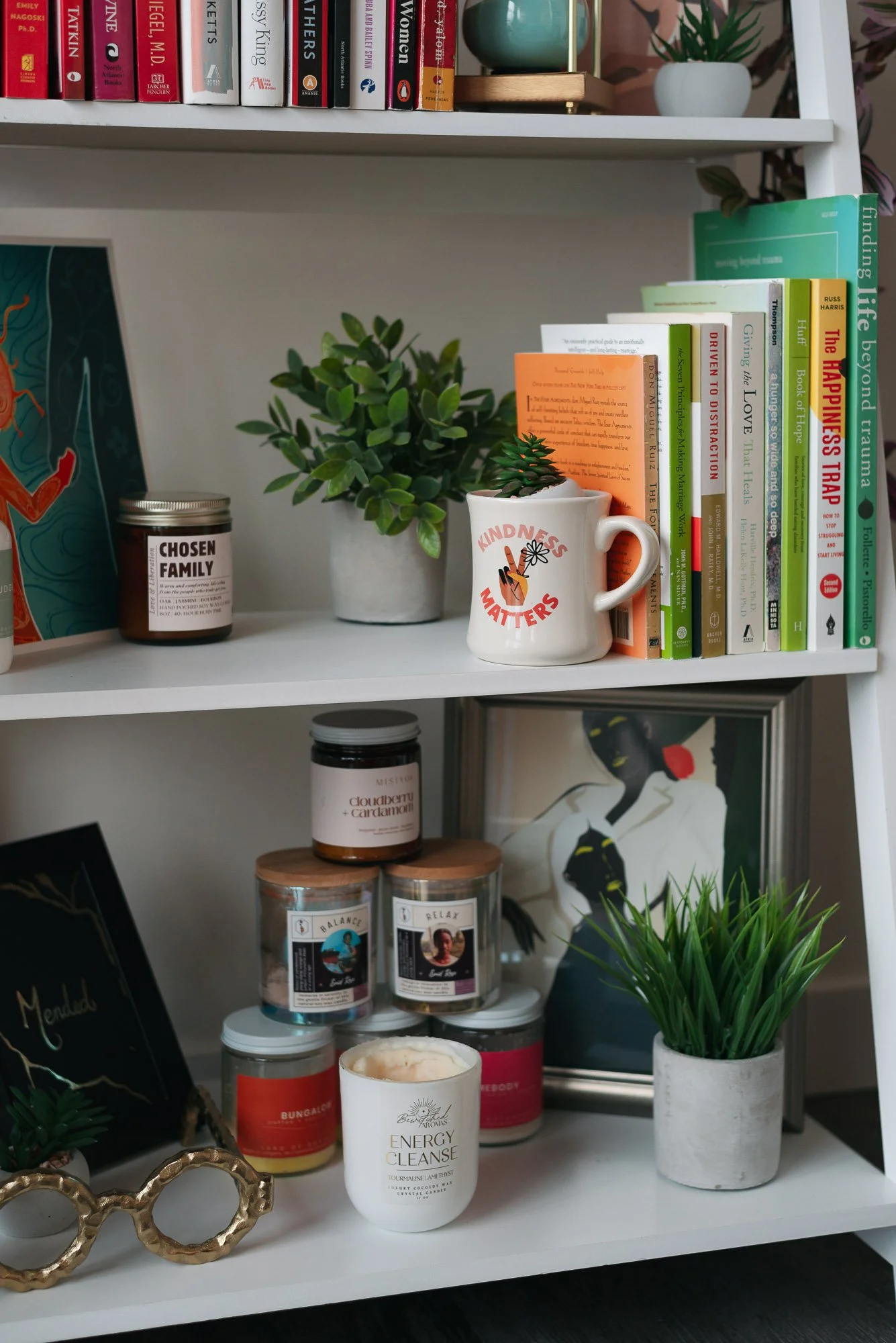 Bookshelf with books, plants, candles, and artwork, including a mug with "Kindness Matters" and a pair of gold glasses.
