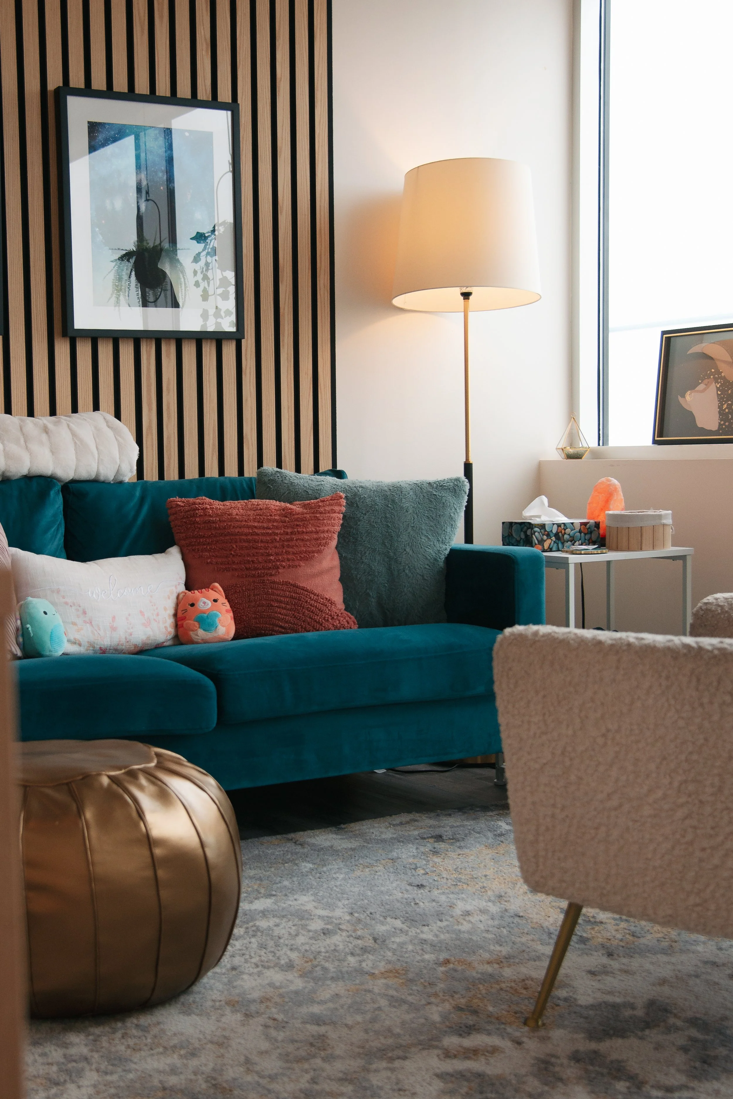 A cozy living room with a teal velvet sofa decorated with various pillows, including a white pillow with an embroidered message, placed on a patterned rug. Next to the sofa is a gold round ottoman. A cream-colored armchair is partially visible in the