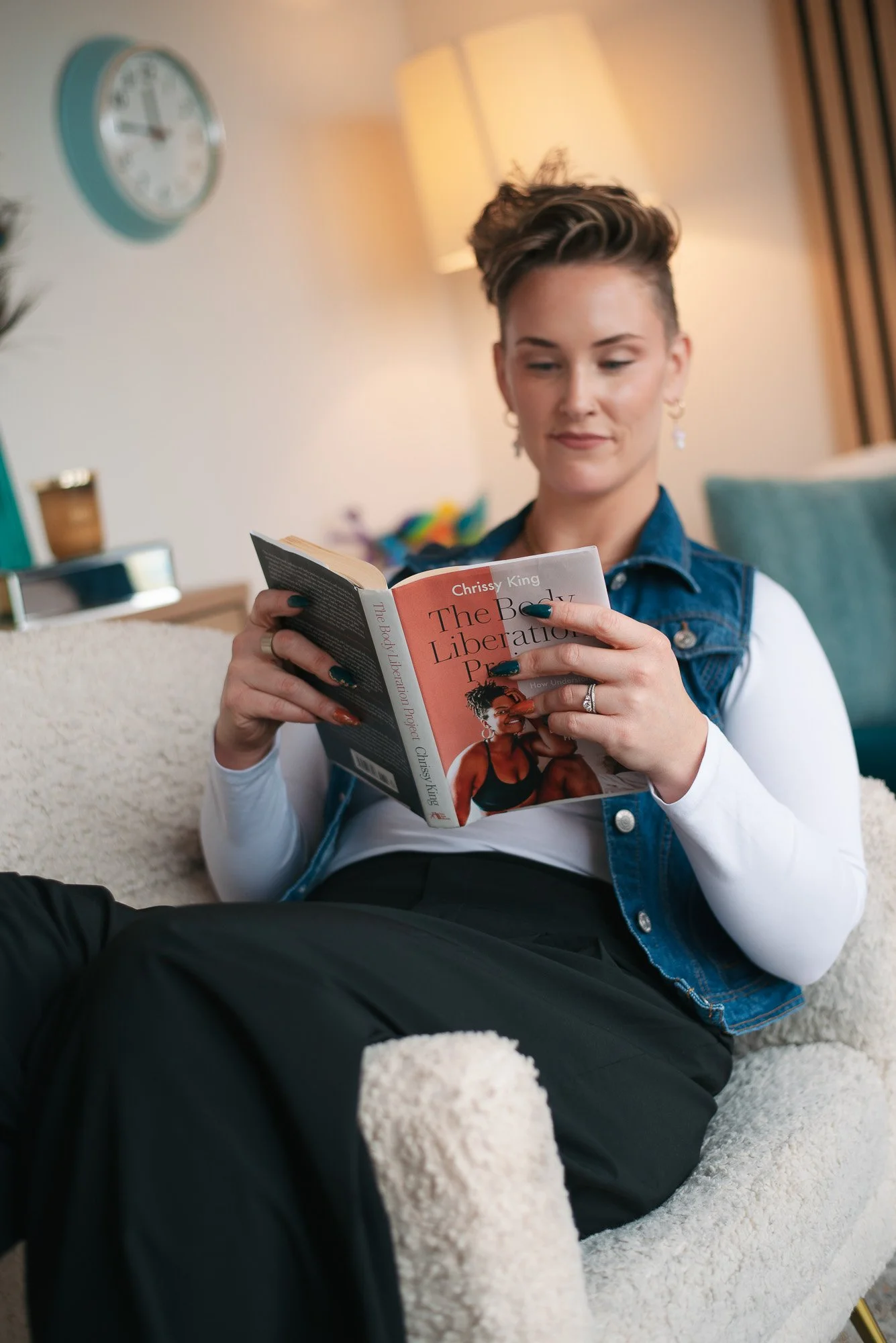 Psychologist in Edmonton with short, styled hair, wearing a white long-sleeve shirt and a sleeveless denim vest, is sitting on a cozy armchair, reading a book titled 'The Body Liberation Project' by Chrissy King in a warmly lit room.
