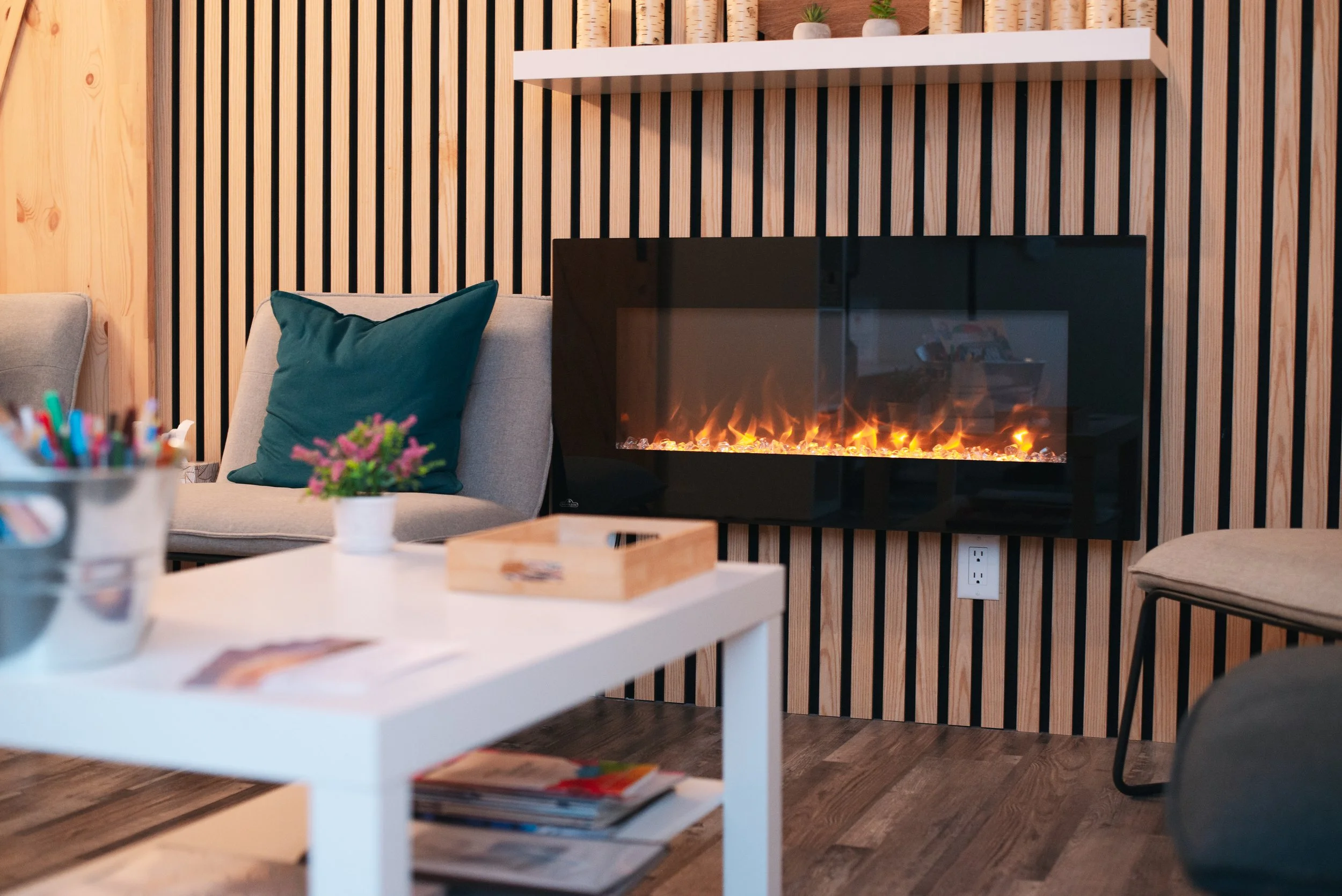 Living room with a wall-mounted electric fireplace with flames visible, a gray sofa with a dark green pillow, a white coffee table with magazines, a small potted plant, and a pencil holder filled with pens and markers.