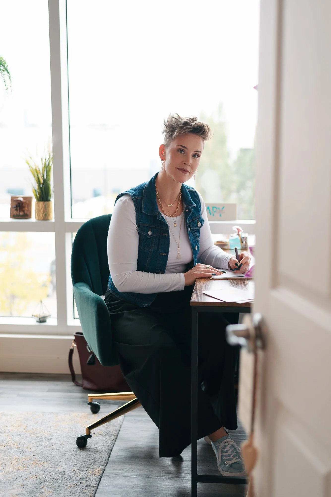 A woman sitting at a desk in a bright office, wearing a denim vest over a white long-sleeve shirt, with short tousled hair and jewelry, holding a pen and smiling at the camera.