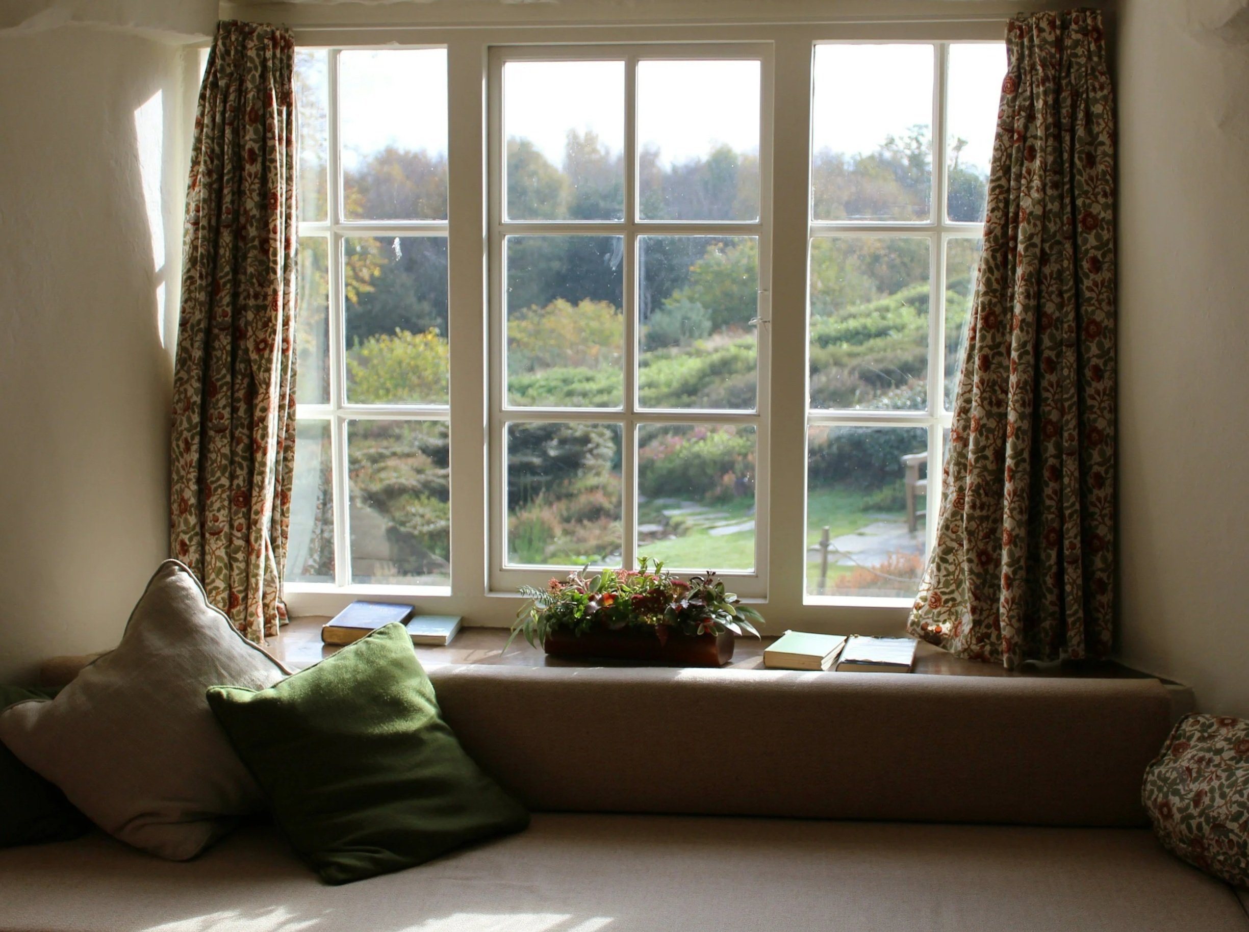 Sunlit window with floral curtains overlooking a garden, with books and a flower arrangement on the window seat, and pillows on a nearby sofa.