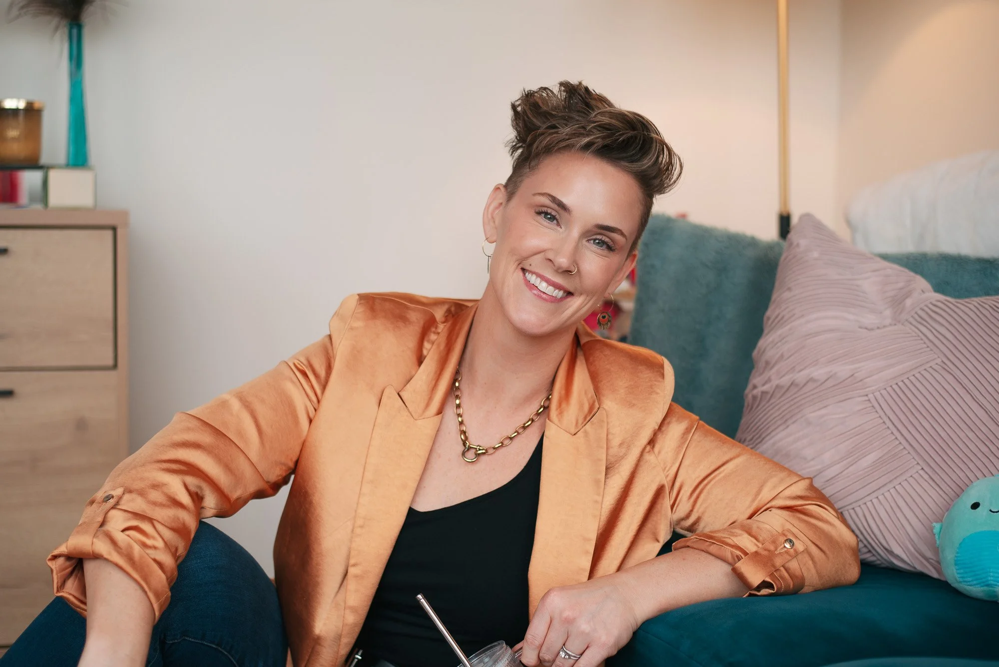 Edmonton therapist portrait, woman with short hair smiling, sitting on a couch with pillows, wearing an orange satin blazer and a black top, holding a smoothie.