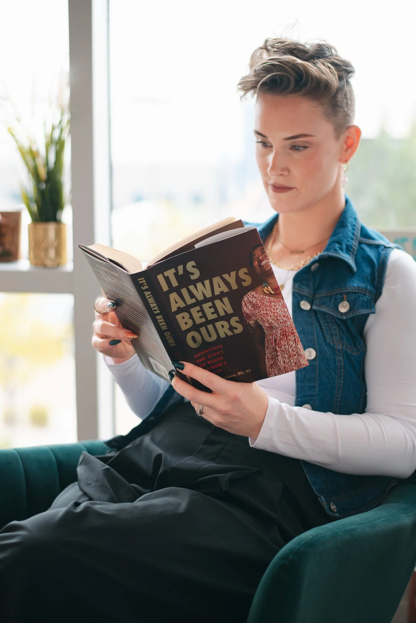 A woman with short hair sitting on a green chair, reading a book titled "It's Always Been Ours" in a bright room with a large window and a potted plant in the background.