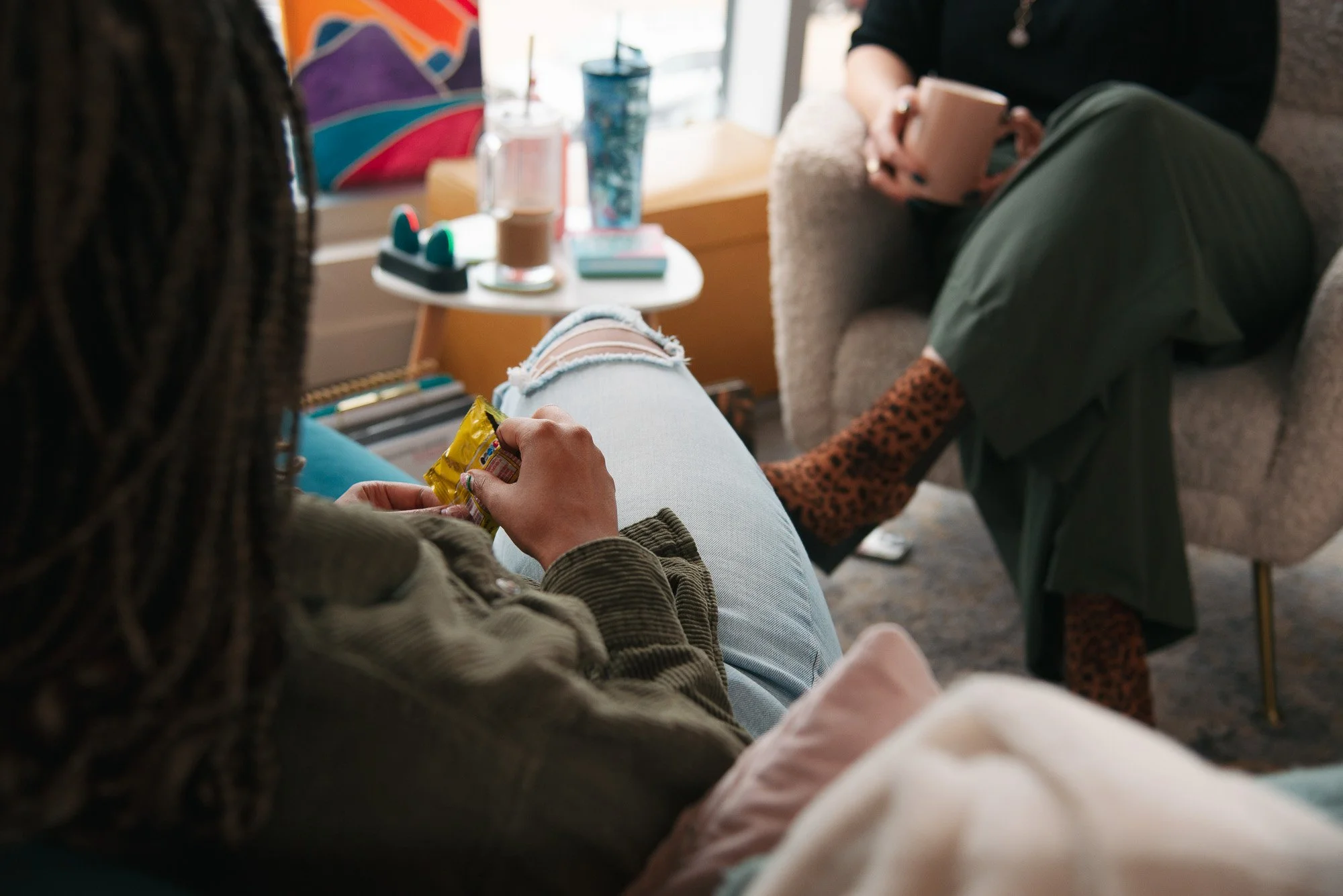Two people sitting and chatting in a cozy indoor space with snacks, drinks, and personal items on a table nearby.