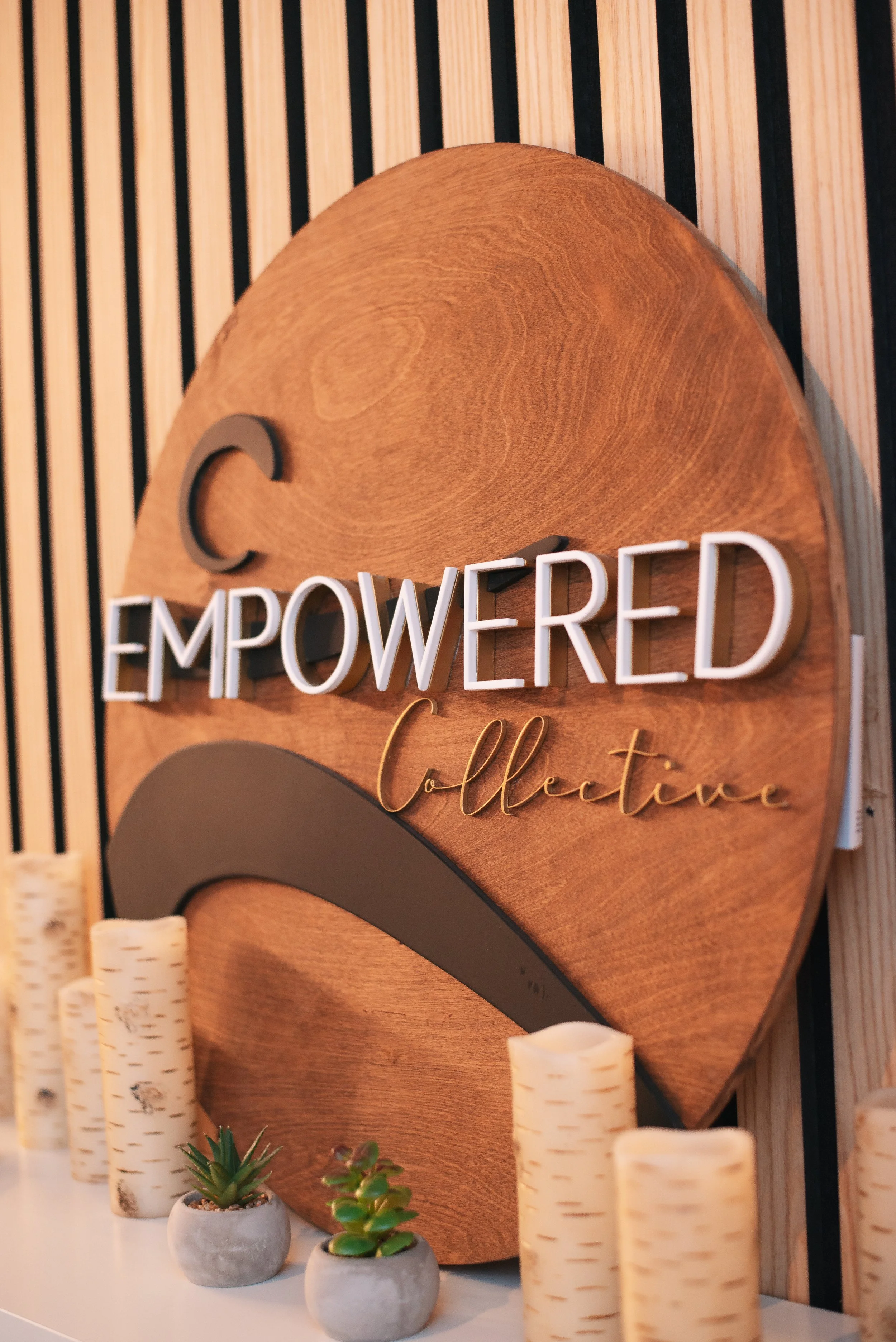 Decorative wooden sign with the words 'Empowered Collection' and a crescent shape, surrounded by small potted succulents and faux birch logs.