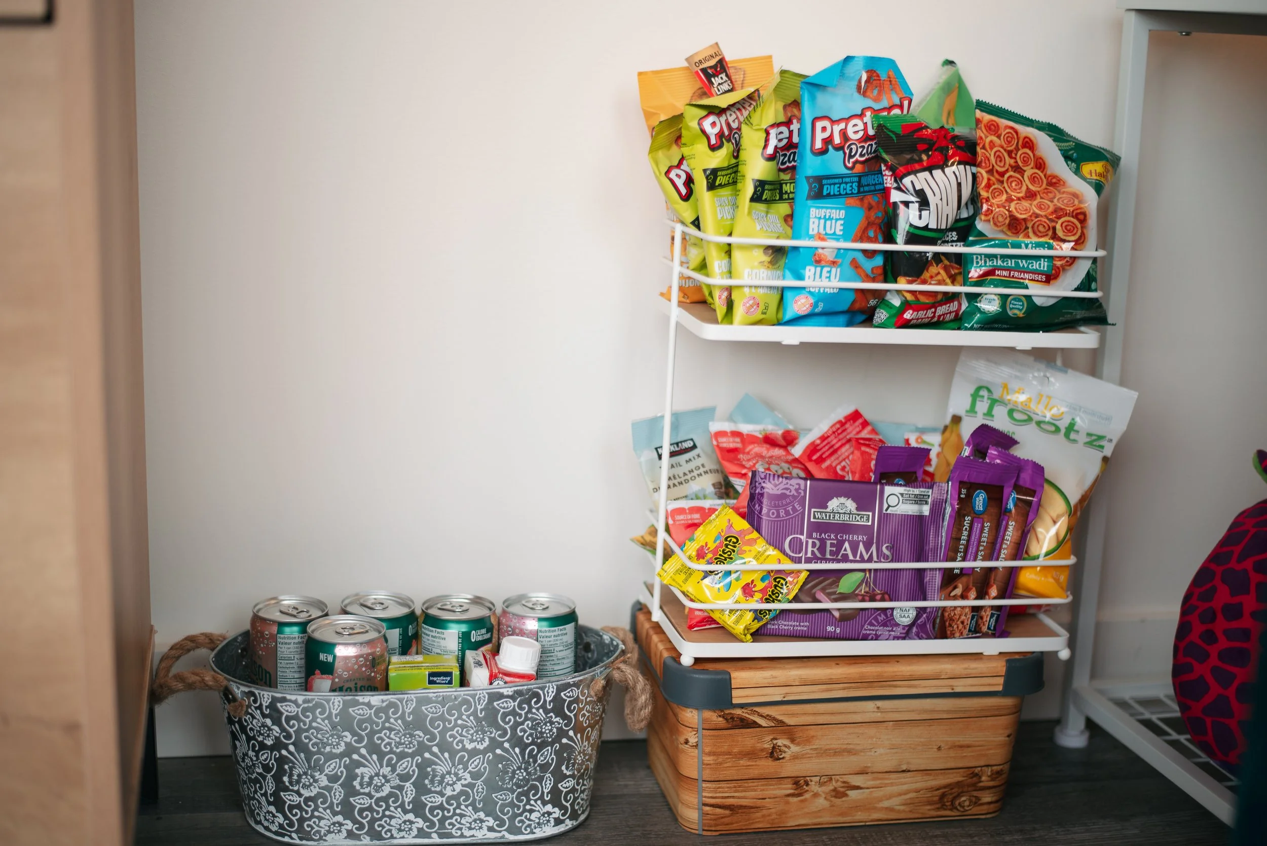 A white metal shelf with snacks, a basket filled with canned drinks, and a wooden storage box on a dark floor in a pantry or storage area.