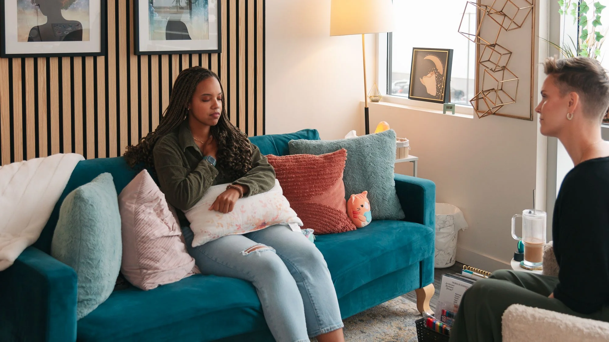 A woman with braided hair sits on a teal sofa with colorful pillows, appearing to be in a counseling session with a therapist in a cozy, decorated room.