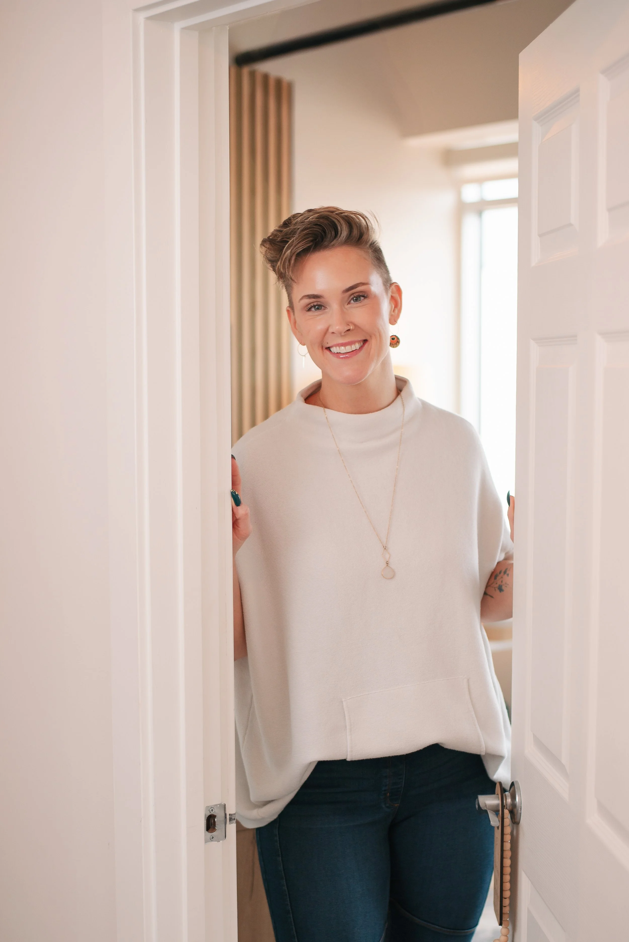A smiling woman with short hair, earrings, and a gold necklace standing at a doorway, wearing a white t-shirt and jeans.