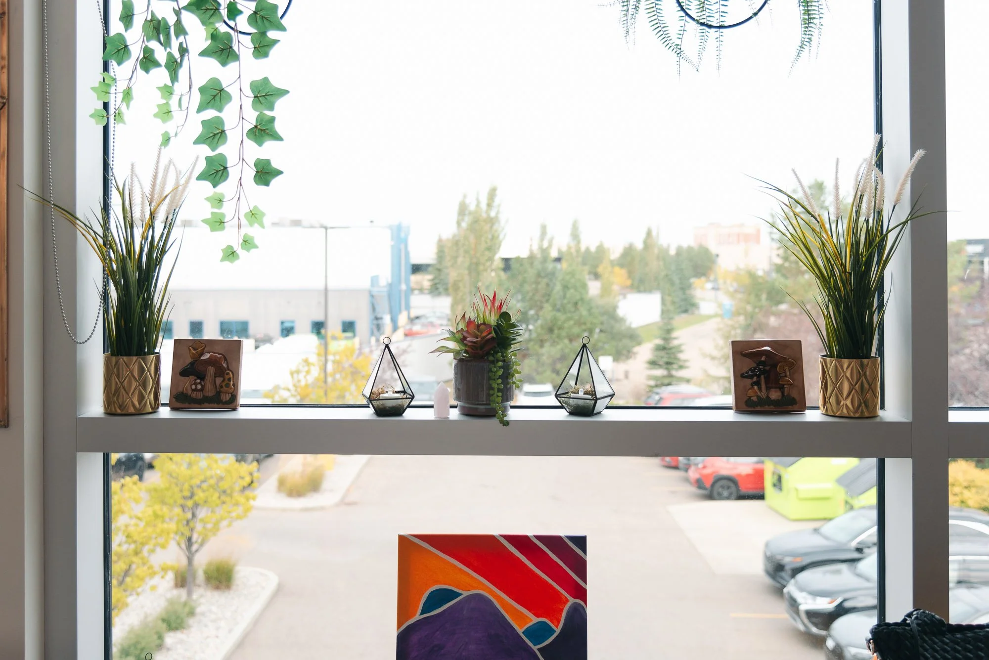 Decorative plants and small sculptures on a windowsill with a view of a parking lot and trees outside.