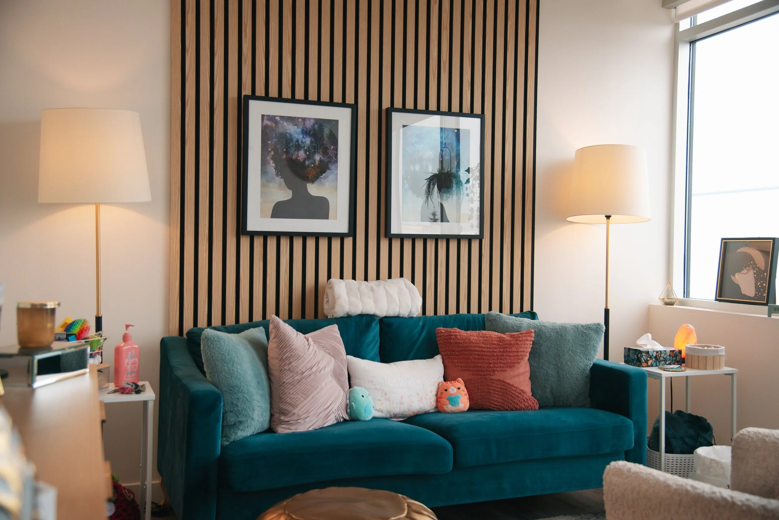 Cozy living room with a teal couch, multiple pillows, framed artwork on a wooden slat wall, and two tall floor lamps, with a window on the right letting in natural light.